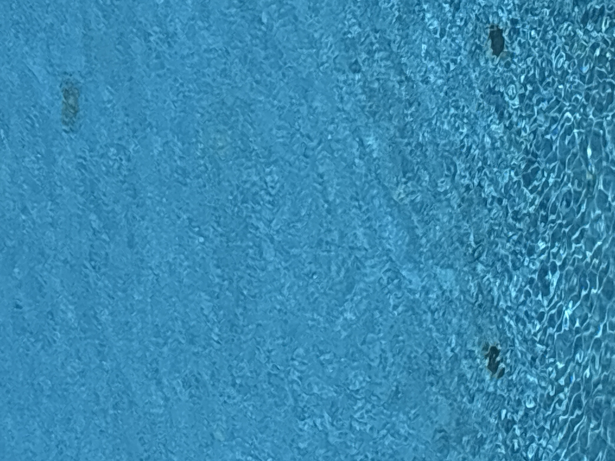 The image displays a close-up view of the rippling surface of a blue swimming pool. The water is agitated, creating patterns of light and shadow on the surface. The lighting suggests it is daytime, with sunlight reflecting off the water. There are no people visible in the frame. The dark shapes near the bottom left and top right appear to be objects submerged in the pool, possibly drains or pool features. The water's color is a vibrant, clear blue, indicative of a clean swimming pool. The image is a vertical shot, focusing solely on the water's texture and movement. The scene could be located at any swimming facility in Varna, Bulgaria, as there are no unique geographical markers to identify a specific location within the city.