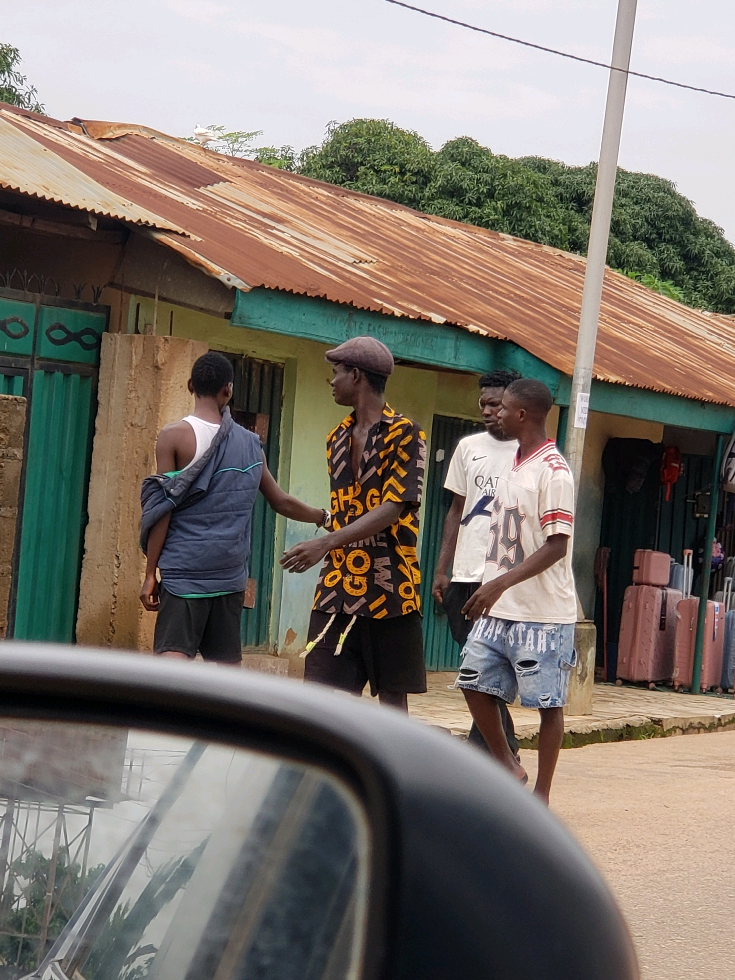 The image depicts a street scene in Narayi, Nigeria, with four young Black men standing in front of a dilapidated building. The building has a rusty corrugated iron roof and appears to be a shop or a dwelling. Green metal gates are visible on the left side of the building. The men are casually dressed. The man on the far left is wearing a blue puffer jacket draped over his shoulder, a white tank top, and black shorts. Next to him, a man in a brown flat cap and a colorful, patterned shirt with orange and black lettering is facing towards the building. To the right, another man is wearing a white t-shirt with "QAT AIR" printed on it. The fourth man, on the far right, is wearing a white jersey with red and white stripes, and distressed denim shorts with "TRAP STAR" written on them in white. He is walking away from the camera. In the background, several suitcases are stacked on the ground near the building. The sky is overcast. The image appears to have been taken from a moving vehicle, as indicated by the reflection in a car's side mirror in the foreground.