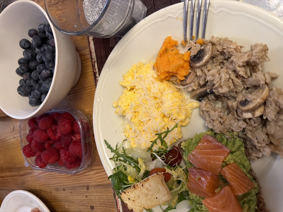 A high-angle, close-up shot captures a meal laid out on a light-colored wooden table in Les Allues, France. The primary focus is a large, light-colored plate holding an array of breakfast or brunch items. On the plate, from left to right, a piece of toast spread with mashed avocado is topped with five distinct cubes of salmon. Adjacent to this is a small mixed green salad containing arugula, cherry tomatoes, and a crouton. Above this, a serving of scrambled eggs is covered with grated cheese. Next to the eggs is a small portion of mashed orange food, likely a vegetable puree, with a silver fork partially inserted into it and an adjacent brown, grain-based dish containing sliced mushrooms and possibly other ingredients like onions.
In the background, positioned on the wooden table, are several other food items and a beverage. A clear plastic container holds a quantity of fresh red raspberries. To its right, a white bowl is filled with numerous dark blue blueberries. Further to the right, a clear glass contains a liquid with many small, dark, circular seeds, consistent with chia seeds in water or a beverage. A sliver of another white plate is visible on the far left edge of the frame. The overall impression is a prepared meal with multiple healthful components.