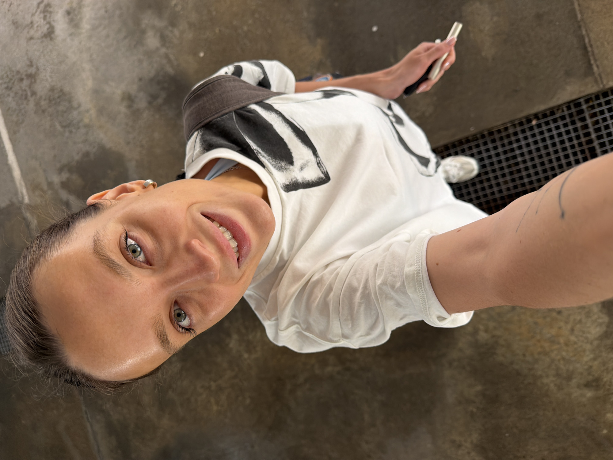 A woman is taking a selfie from a high angle, looking up at the camera. She has light skin, green eyes, and her dark hair is pulled back. She is wearing a white t-shirt with a black abstract print and a dark backpack. She is smiling. Her left arm is extended towards the camera, holding what appears to be a phone. Her right hand is visible holding a dark object, possibly a phone or wallet. She is wearing white sneakers. The background appears to be a textured, wet concrete or asphalt surface, possibly a street or walkway. A metal grate is visible on the ground in front of her. The overall impression is of an outdoor urban setting. The provided crops help to focus on the woman's face, her clothing, and the immediate surroundings. While the image itself does not contain any definitive landmarks or signs to identify the specific location, if this image were to be associated with Budva, Montenegro, the environment could be interpreted as a public space within the city, such as a promenade, square, or street.
