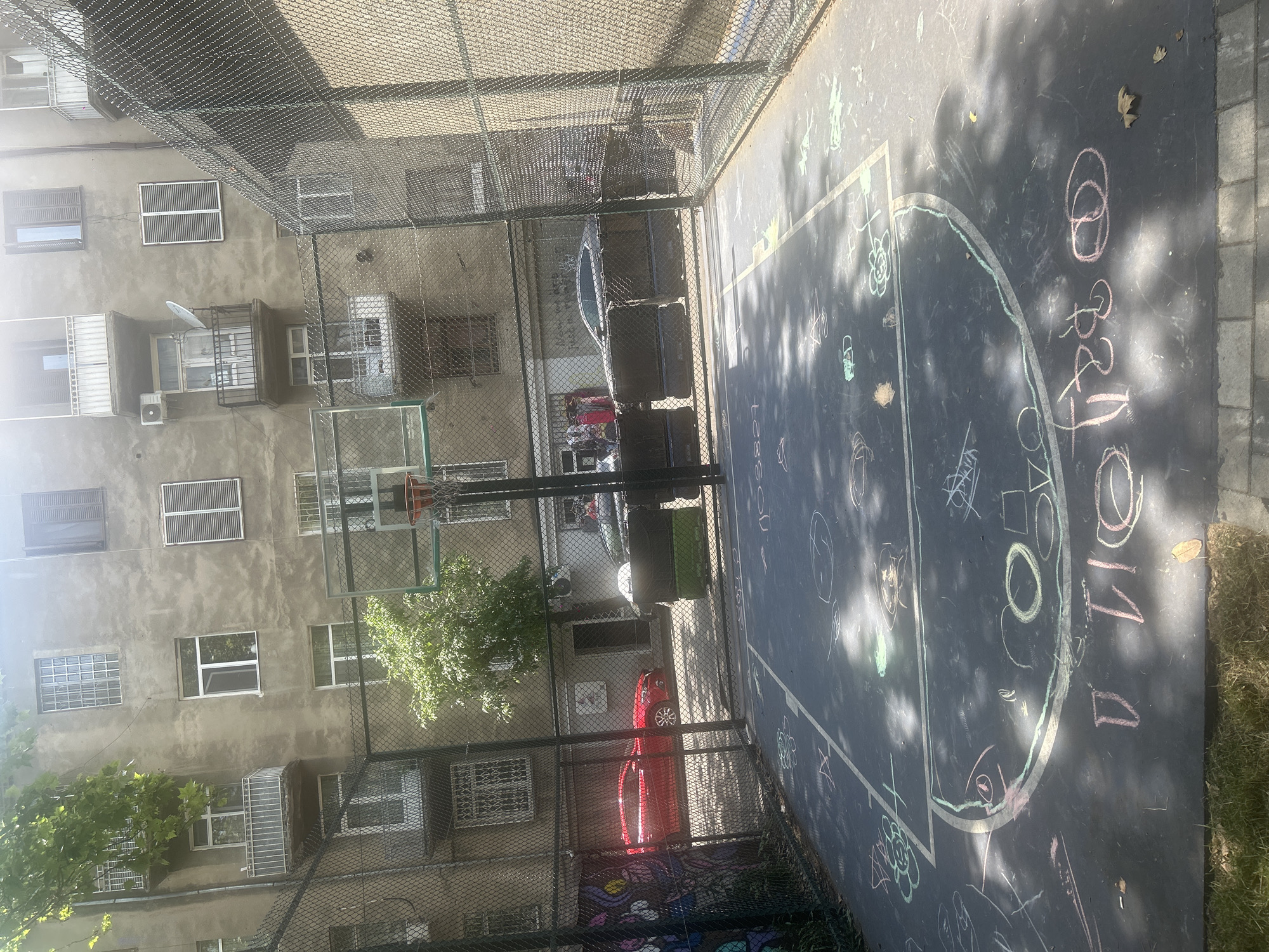 The image shows an outdoor basketball court in an urban environment, likely in Tbilisi, Georgia. The court surface is black and covered with various chalk drawings, including shapes, flowers, and text. A green chain-link fence encloses the court. Behind the fence, a weathered, multi-story building with numerous windows and balconies is visible. Trees and some vehicles, including a red car, are also present in the background. The scene appears to be illuminated by sunlight, casting shadows on the court.