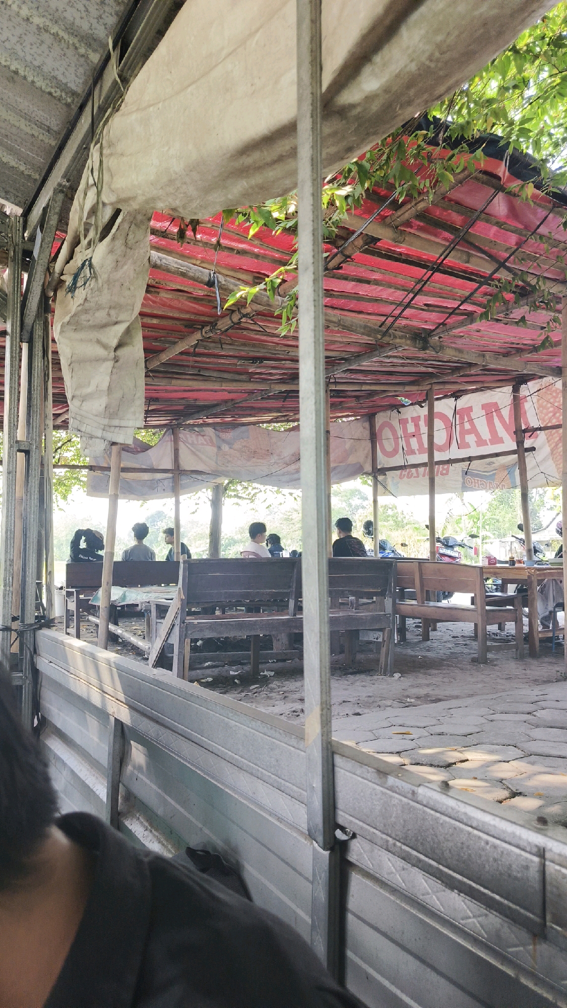 A daytime, medium shot captures an outdoor seating area of a food stall or restaurant in Ngemplak, Indonesia. The foreground is dominated by the side of a vehicle, possibly a truck bed, with corrugated metal siding. Beyond this, wooden benches and tables are arranged under a makeshift roof. The roof is constructed from bamboo poles supporting a red tarp and a lighter, aged fabric. Green foliage from overhanging trees is visible. Several people are seated at the tables, their backs mostly to the camera, suggesting they are customers. A banner with Indonesian text, partially obscured, hangs in the background. The ground appears to be paved with hexagonal tiles and dirt. The overall impression is a casual, open-air dining setting.