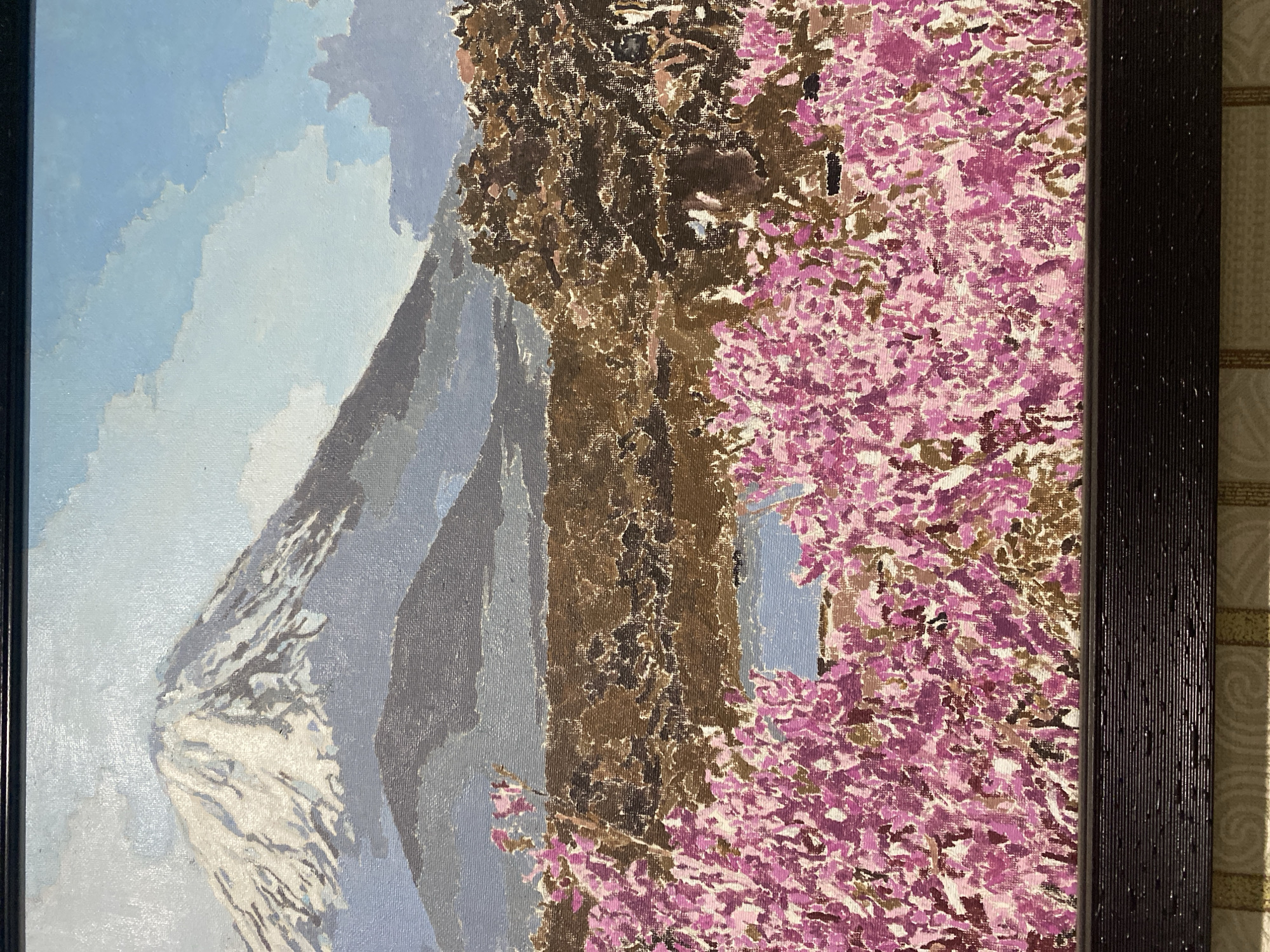 This is a painting depicting a landscape with Mount Fuji in the background. In the foreground, there are clusters of pink flowers, and behind them, there is a body of water surrounded by trees. The sky is blue with white clouds. The painting is framed by a dark, textured frame. There is no indication of the city of Chernogorsk, Russia, within the image.