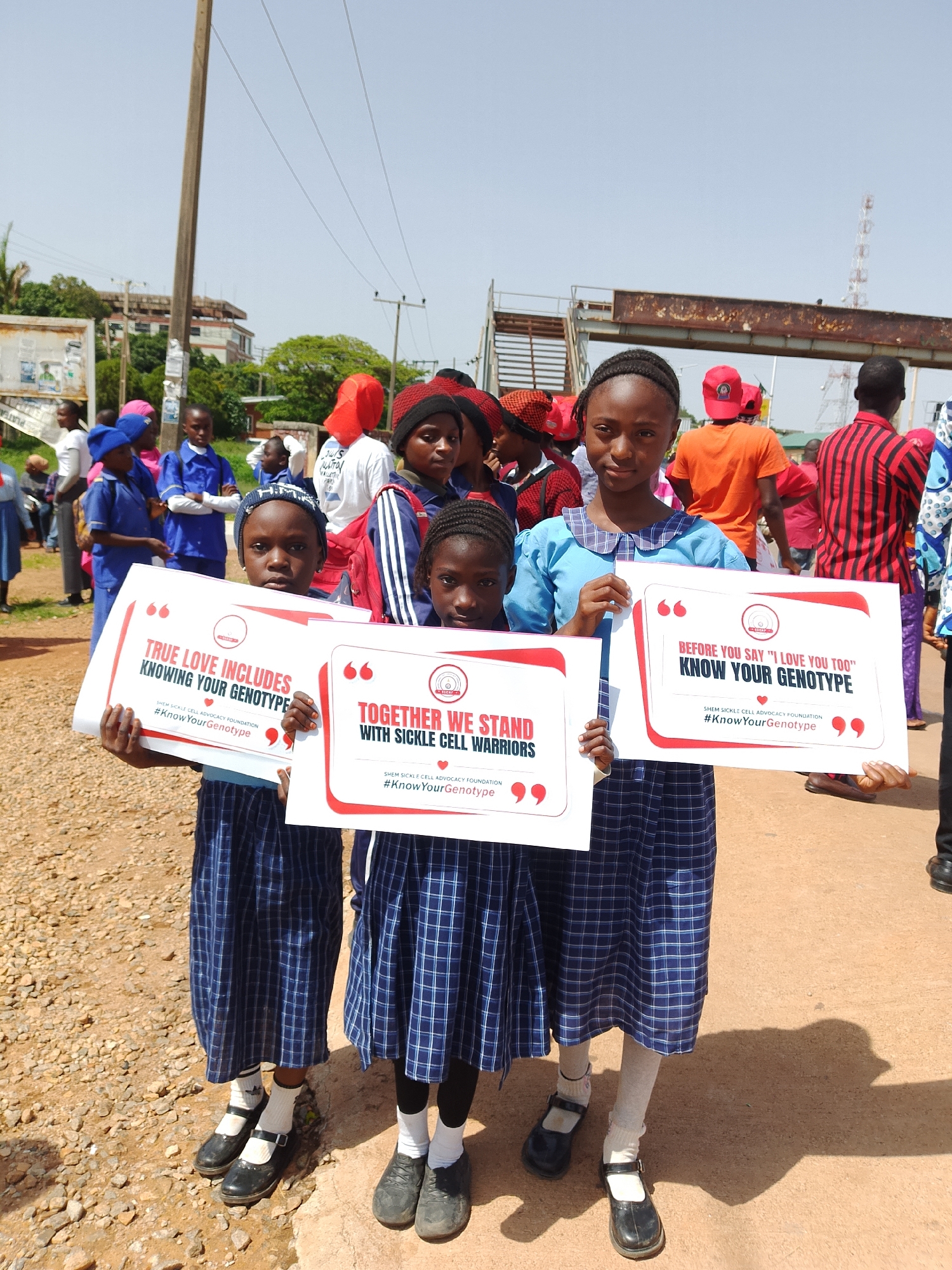 In Jos, Nigeria, three young girls in school uniforms stand holding signs. The girl in the center wears a blue plaid skirt and a light blue shirt, holding a sign that reads "TRUE LOVE INCLUDES KNOWING YOUR GENOTYPE". The girl to her right wears a similar uniform and holds a sign that says, "TOGETHER WE STAND WITH SICKLE CELL WARRIORS". The girl on the far right, partially visible, is also in uniform and holds a sign that states, "BEFORE YOU SAY 'I LOVE YOU TOO' KNOW YOUR GENOTYPE". Behind them, a crowd of people, many wearing red, are gathered. The environment appears to be outdoors, possibly on a street or open area, with a dusty ground and a few trees and buildings visible in the background. A utility pole is prominent to the left of the frame, and an elevated walkway or bridge structure is visible in the distance. The sky is clear and bright.