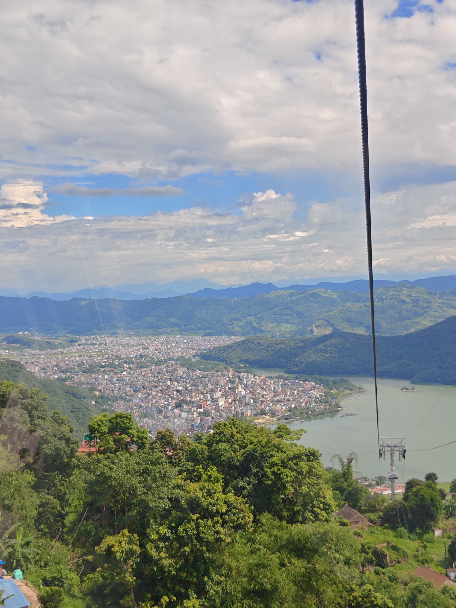 This image captures a panoramic view of Pokhara, Nepal, taken from an elevated vantage point, likely during a cable car ride, given the prominent cable line running down the right side of the frame. The city of Pokhara sprawls across the landscape, characterized by a dense concentration of buildings with predominantly light-colored roofs, interspersed with greenery. A large body of water, likely Phewa Lake, is visible to the right of the city, with a few boats dotting its surface. The lake is bordered by lush, green hills that recede into the distance, forming multiple layers of mountains under a cloudy sky. Sunlight filters through gaps in the clouds, casting dappled light and shadows across the scene. The foreground features dense green foliage, with trees and vegetation obscuring parts of the lower section of the image. A cable car station or pylon is visible in the lower right quadrant, further indicating the perspective. There are no visible people or animals in the frame.