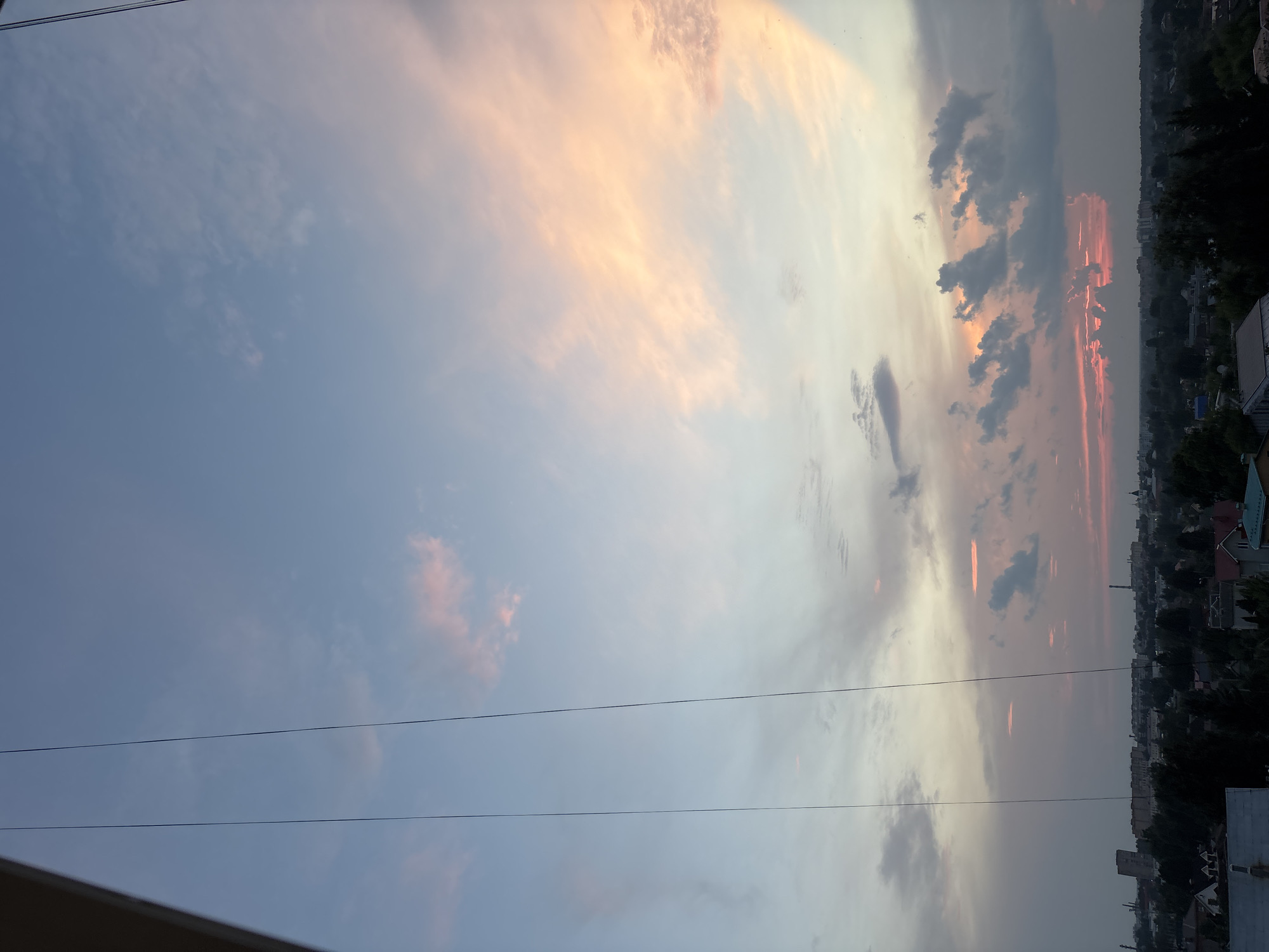 The image shows a view of the sky above the city of Klin, Russia, during what appears to be sunset or twilight. The sky is filled with clouds, with streaks of pink and orange light illuminating them from the setting sun. The lower portion of the image shows a dense, green tree line and the rooftops of various buildings, including residential houses and some taller structures that might be industrial or commercial. Two thin, dark wires are visible in the foreground, stretching vertically across the frame, suggesting the viewpoint is from a window or balcony. The overall atmosphere is serene and somewhat dramatic due to the colorful sky.