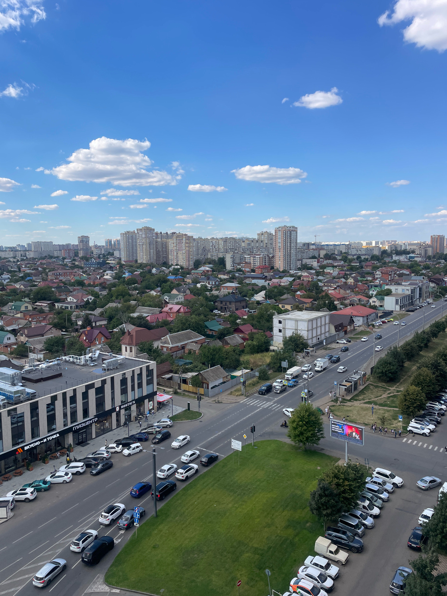 The aerial view depicts a bustling urban scene in Krasnodar, Russia, under a bright blue sky dotted with fluffy white clouds. A multi-lane road, featuring both moving traffic and parked vehicles, dominates the foreground and midground. On the left side of the road, a modern commercial building with glass windows and striped detailing is visible, bearing the signage "ГЛАЗЦЕНТР" and "ВКУСНО — И ТОЧКА". Across the street, numerous residential houses with red and brown rooftops are nestled amongst lush green trees, giving way to a dense cityscape of high-rise apartment buildings in the background. Cars of various models and colors are seen on the road and in parking areas. A large billboard with a red percentage sign and Russian text is also present. The overall impression is one of a well-populated and active urban environment.