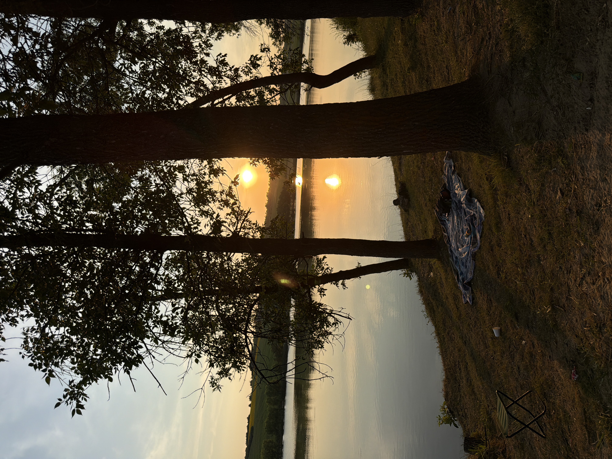 This is a horizontal, medium-shot, eye-level photograph taken outdoors at sunset. The scene depicts a tranquil lakeside setting with silhouetted trees in the foreground and a calm body of water reflecting the warm hues of the setting sun. The sky is a gradient of pale blue and soft yellow, with a few wispy clouds. The sun, a bright orange orb, is partially obscured by trees in the mid-ground, casting a golden glow on the water.
On the grassy bank in the foreground, there is a folded green camping chair on the left. Further to the right, a blue and white patterned blanket is spread out on the ground, with some items on it that are difficult to discern. A white paper cup is visible on the ground near the blanket. The ground is covered in dry grass and some sandy patches. The trees are tall and slender with dark bark and dense foliage, their leaves forming dark silhouettes against the bright sky. The overall impression is one of peaceful solitude and natural beauty. The location is Găleștii Noi, Moldova.