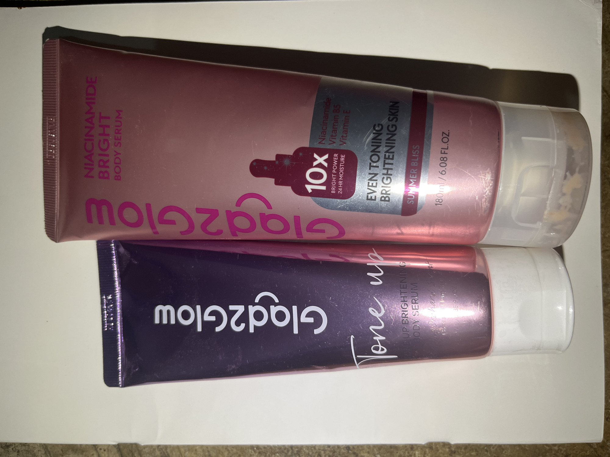 Two tubes of Glad2Glow body serum are placed side-by-side against a white background, with subtle shadow cast to the right of the objects. The tube on the left is purple with white text and reads "Glad2Glow Tone up" and "TONE UP BRIGHTENING BODY SERUM". The tube on the right is pink with purple text and reads "Glad2Glow" and "NIACINAMIDE BRIGHT BODY SERUM". A label on the pink tube indicates "10x BRIGHT POWER 24 HR MOISTURE" along with "Niacinamide, Vitamin B5, Vitamin E" and "EVEN TONING BRIGHTENING SKIN SUMMER BLISS 180ml / 6.08 FL.OZ.". The image does not contain any information about the city of Meliau Hulu, Indonesia.