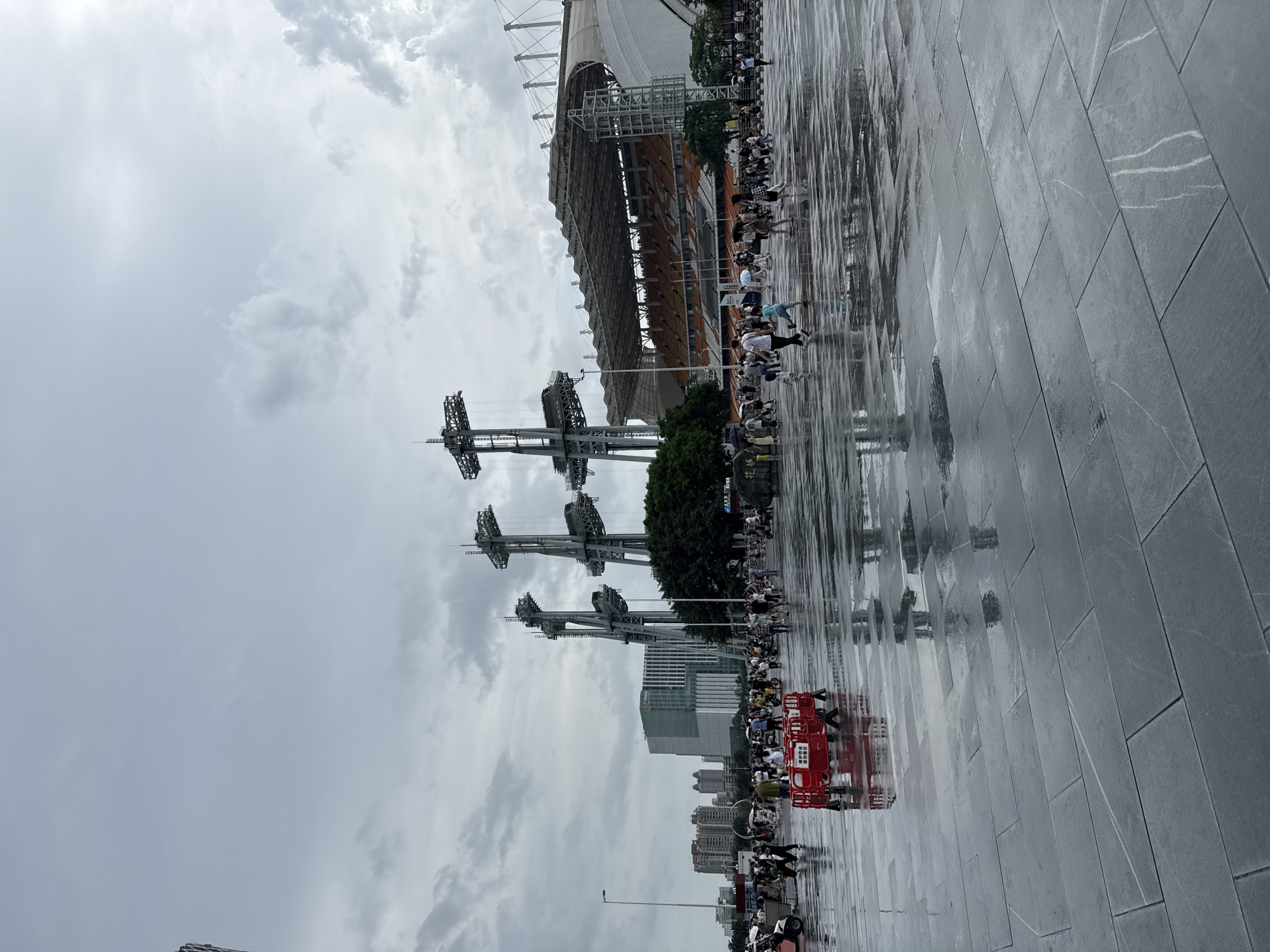 The image depicts an outdoor plaza in Tianhe District, China, on a cloudy day with wet pavement. A large stadium with orange seating and a white roof is visible in the background. Several tall, industrial-looking structures, possibly part of the stadium's lighting or support system, stand prominently in the foreground and midground.
A diverse crowd of people is present, many walking across the wet plaza. Some individuals are holding umbrellas, suggesting recent or ongoing rain. A red, portable barrier with Chinese text is positioned in the midground. The pavement, made of large, rectangular stone tiles, reflects the sky and surrounding structures due to the accumulated water. In the distance, several modern buildings and trees contribute to the urban landscape. The overall scene suggests a public gathering space, likely near a sports or entertainment venue, experiencing overcast weather conditions.