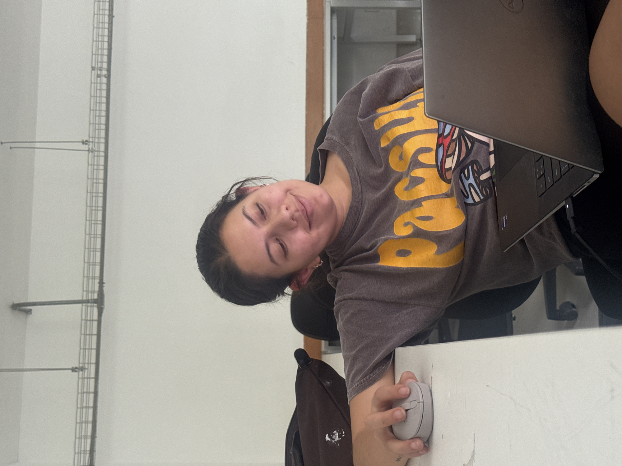 The image depicts a young woman indoors. She is seated at a white desk, holding a computer mouse with her right hand. A gray Dell laptop rests on the desk in front of her. She is wearing a gray t-shirt with yellow lettering and a design featuring cartoon characters. A brown backpack is visible near her. The background includes a wall with a metal support structure and a glimpse of a window or opening with a white interior. Without any specific contextual clues in the image, I can not confirm the specific location in San Andrés Cholula, Mexico, but this appears to be a study or work environment.