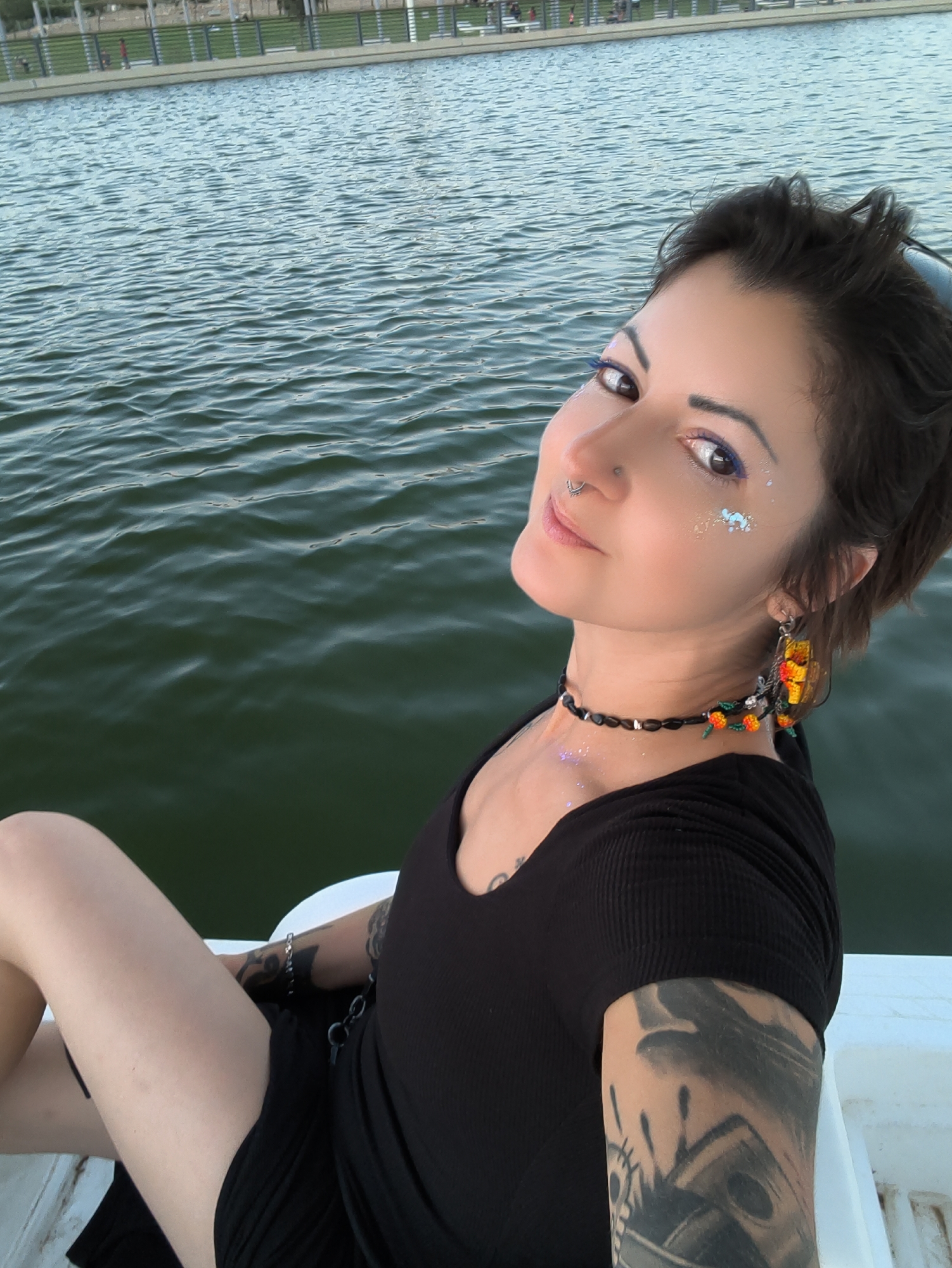 The image shows a young woman sitting in a white pedal boat on a body of water. She is wearing a black, ribbed, short-sleeved shirt and a black skirt. She has tattoos on her arms and a nose ring. Her makeup includes blue eyeliner and glitter on her cheeks. She is also wearing a beaded necklace and large, colorful earrings. The water is calm with ripples, and in the background, there is a grassy area with a fence and people visible. The scene appears to be a park or recreational area. It is possible this is the lake in Be'er Sheva, Israel.