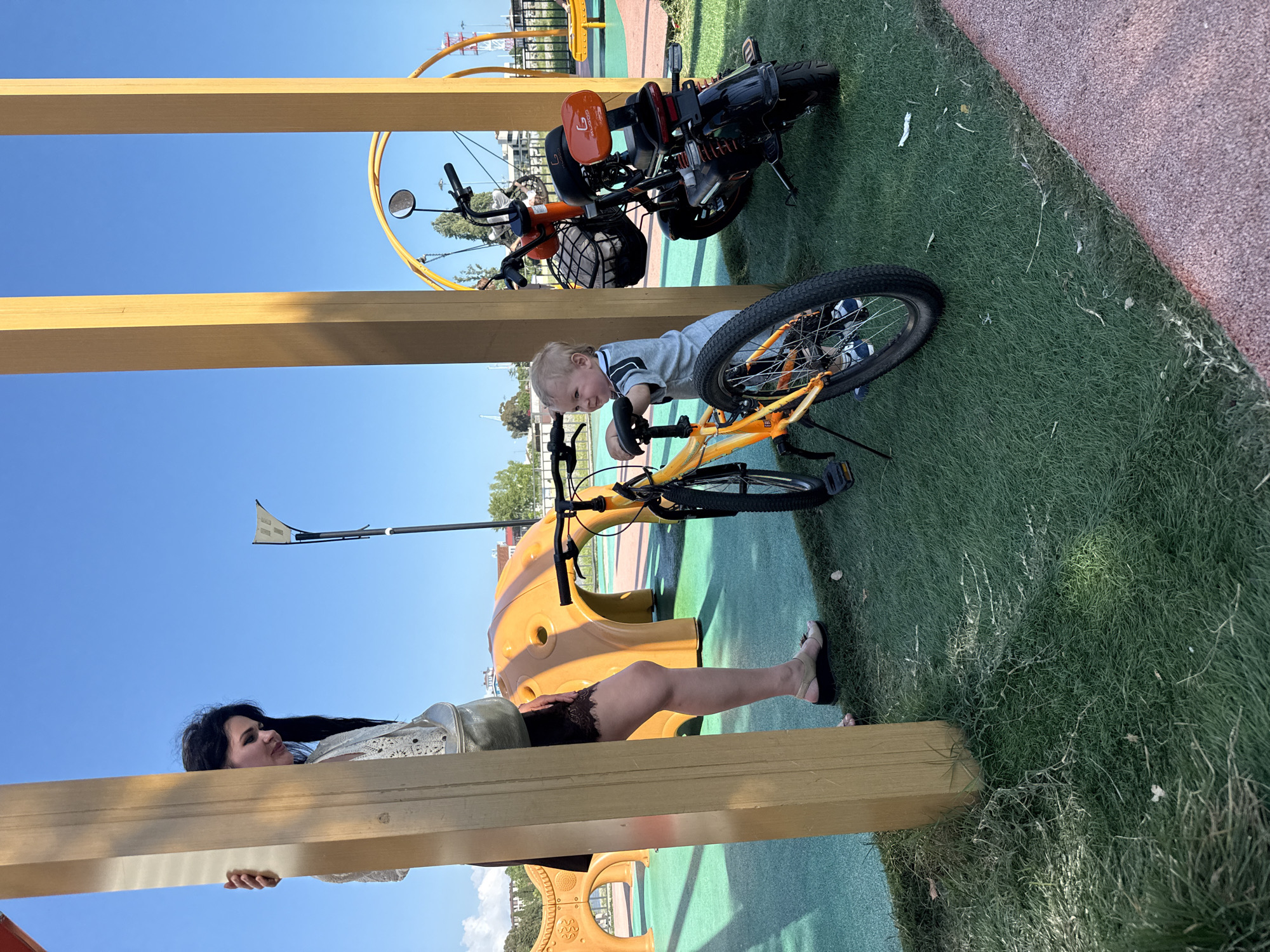 In Sochi, Russia, a woman stands partially obscured by a wooden pillar, her hand resting on it. She is wearing a white open-knit top and a black skirt. In front of her, a young child is seated on a yellow bicycle, looking to the left. Behind the child, a black and orange electric scooter is parked. The scene is set on a playground with green turf and a yellow climbing structure in the background. The sky is clear and blue, indicating a sunny day.