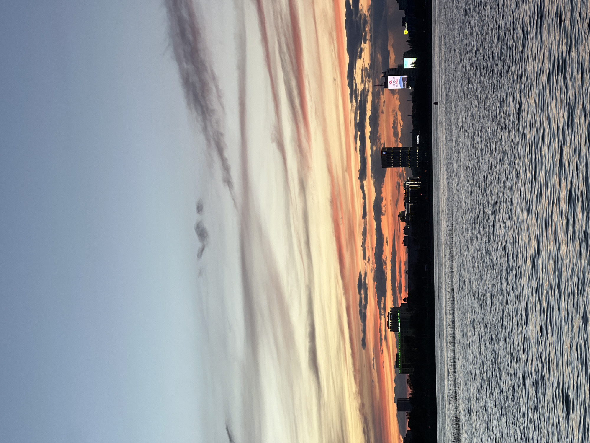 The image depicts a cityscape at sunset, seemingly in Kazan, Russia, based on architectural cues and landmarks. The right third of the image is a body of water with wavy surface, reflecting light from the sky. The city skyline is a dark silhouette against the vibrant colors of the sunset. There are buildings of varying heights, including some high-rises. Two prominent advertising billboards appear on the skyline. The sky features a gradient from light blue to orange and red with dark clouds forming vertical patterns. The image is rotated 90 degrees counter-clockwise.