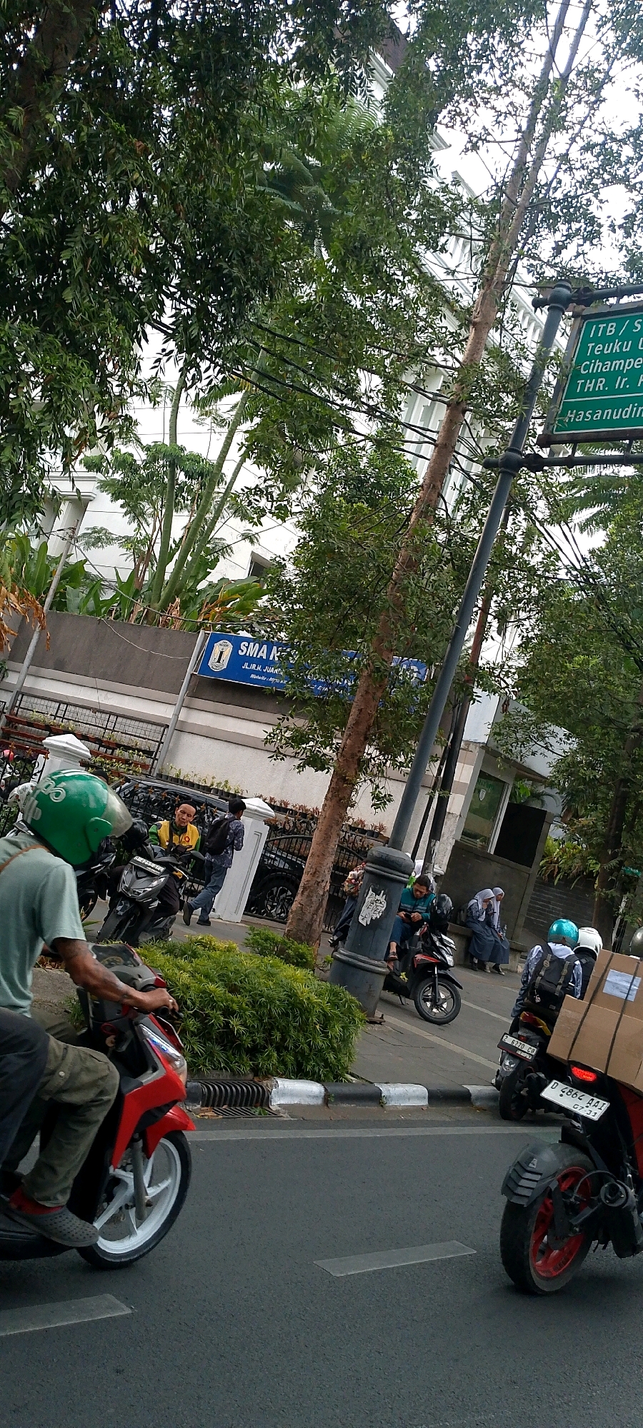 The image depicts a street scene in Bandung City, Indonesia, with a focus on motorbikes and pedestrians on a sidewalk next to a road. A person wearing a green helmet is riding a red motorbike in the foreground. Further back, several other motorbikes are parked or in motion. On the sidewalk, individuals are seen walking, sitting, and interacting. Behind the sidewalk, there is a building with a sign that reads "SMA NEGERI". A street sign is also visible, indicating directions and distances, with text that appears to be in Indonesian. Lush green trees and foliage line the street, and overhead power lines are present. The overall atmosphere suggests a busy urban environment.