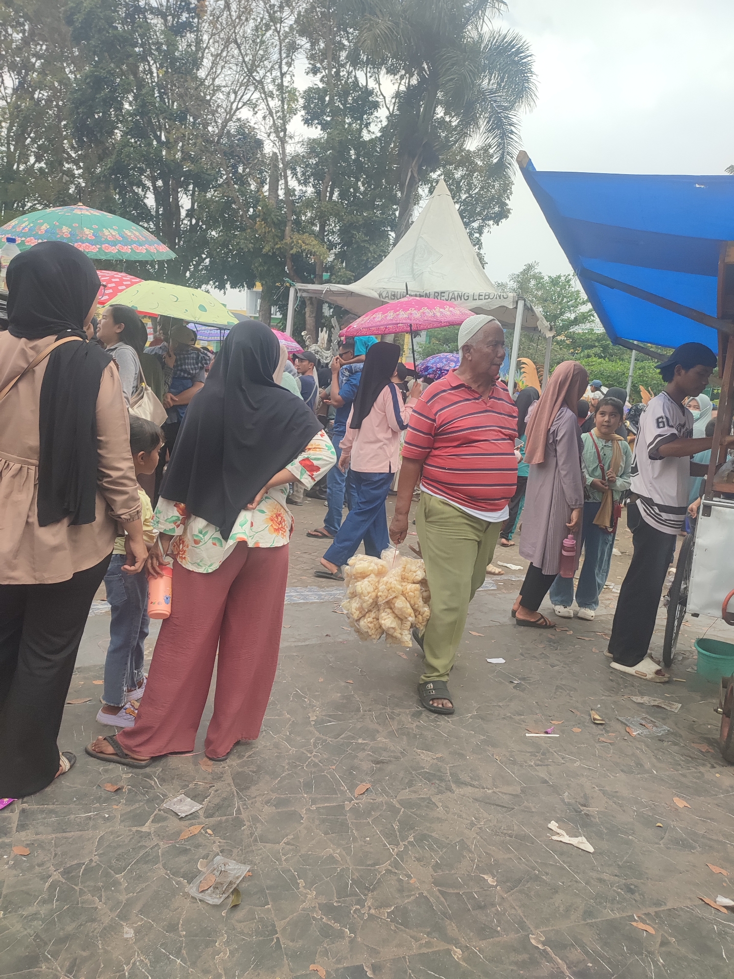 This image depicts an outdoor scene in Dwi Tunggal, Indonesia, likely a market or public gathering area. A group of people are present, some walking, others standing. In the foreground, a man in a red and white striped shirt and green pants walks towards the right, carrying a large bag of puffed snacks. To his left, a woman wearing a black hijab and a floral-patterned top with rust-colored pants is visible from the back, facing left. Further left, another woman in a black hijab and a light brown top is seen from the back, also facing left, holding a small child's hand. Several umbrellas, some patterned and some plain, are scattered throughout the scene, suggesting it might be a partly cloudy or sunny day, or that people are seeking shade. In the background, white and blue tents are visible, along with trees and other structures. The ground appears to be paved with large, dark tiles. The overall atmosphere suggests a busy, communal event.