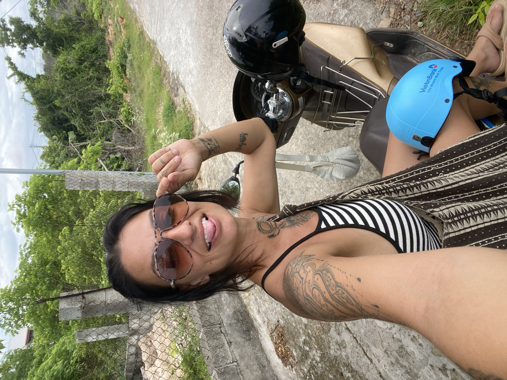 A woman with dark hair, sunglasses, and a black and white striped top sits on a scooter in Mũi Né, Vietnam. She is sticking her tongue out and touching her sunglasses with her right hand. A black helmet is on the back of the scooter, and a light blue helmet with a VietinBank logo is on her lap. The background shows a dirt road, overgrown vegetation, and a fence.