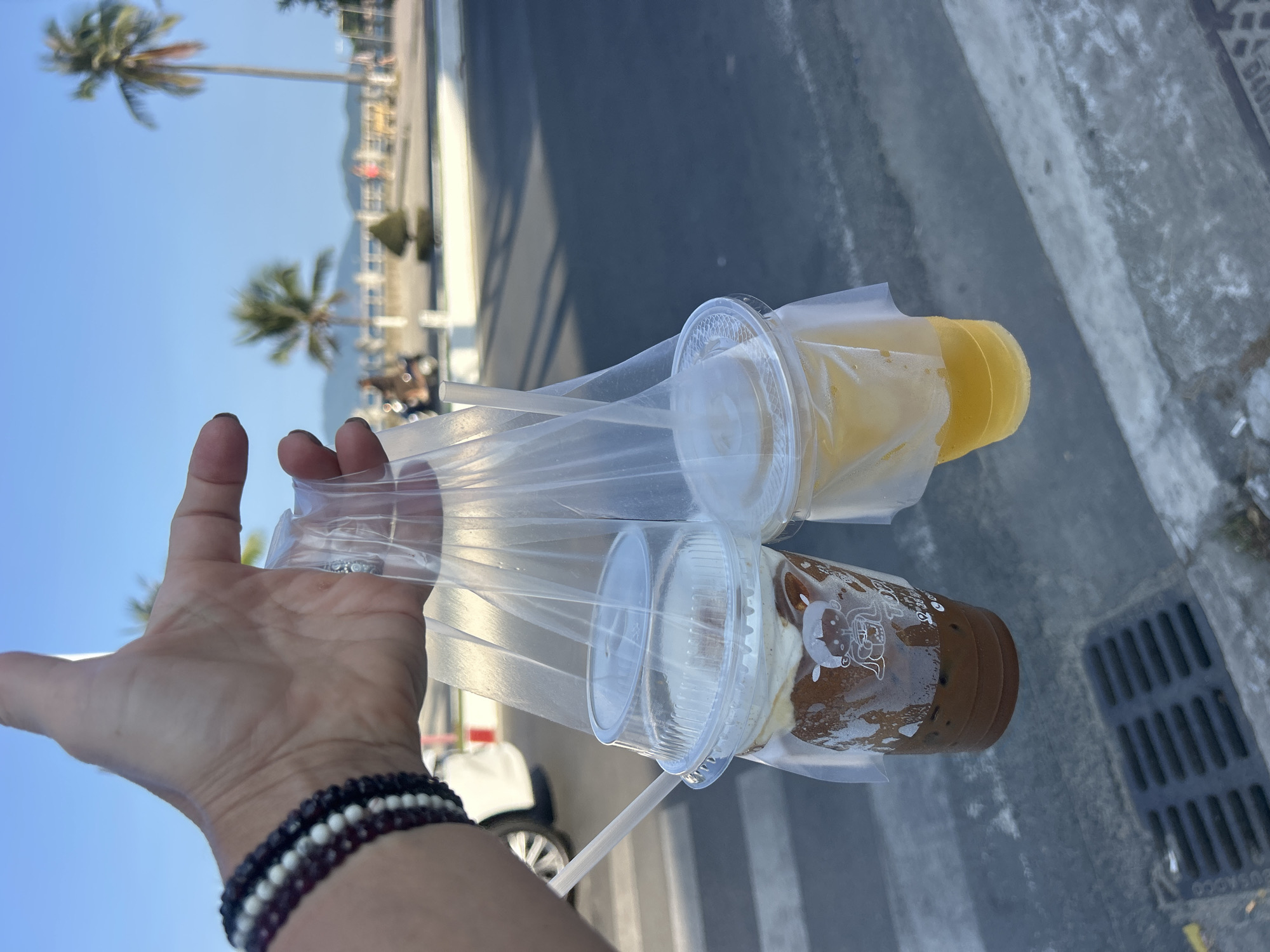A first-person shot captures a hand holding two beverages in a clear plastic carrier bag, likely along a coastal promenade or street in Bắc Nha Trang, Vietnam. The hand, adorned with stacked beaded bracelets (dark purple/black and white), firmly grips the plastic holder. One drink is a vibrant yellow or orange, while the other is a dark brown, possibly chocolate or coffee, topped with a layer of white foam or cream, and features an unidentifiable white illustration or text on its plastic cup. A plastic straw protrudes from one of the drinks.
The setting is outdoors on a clear, sunny day, indicated by the bright blue sky. In the background, tall palm trees stand against the sky, suggesting a tropical or subtropical environment. Beyond a road or path, a body of water (likely the sea or ocean) stretches to the horizon, meeting distant mountains or hills. The foreground features a concrete curb and a metal storm drain or grate on the ground. A white vehicle is partially visible further down the road. The overall scene evokes a leisurely moment during pleasant weather, perhaps during a vacation. No explicit text is clearly legible on the cups or the storm drain.