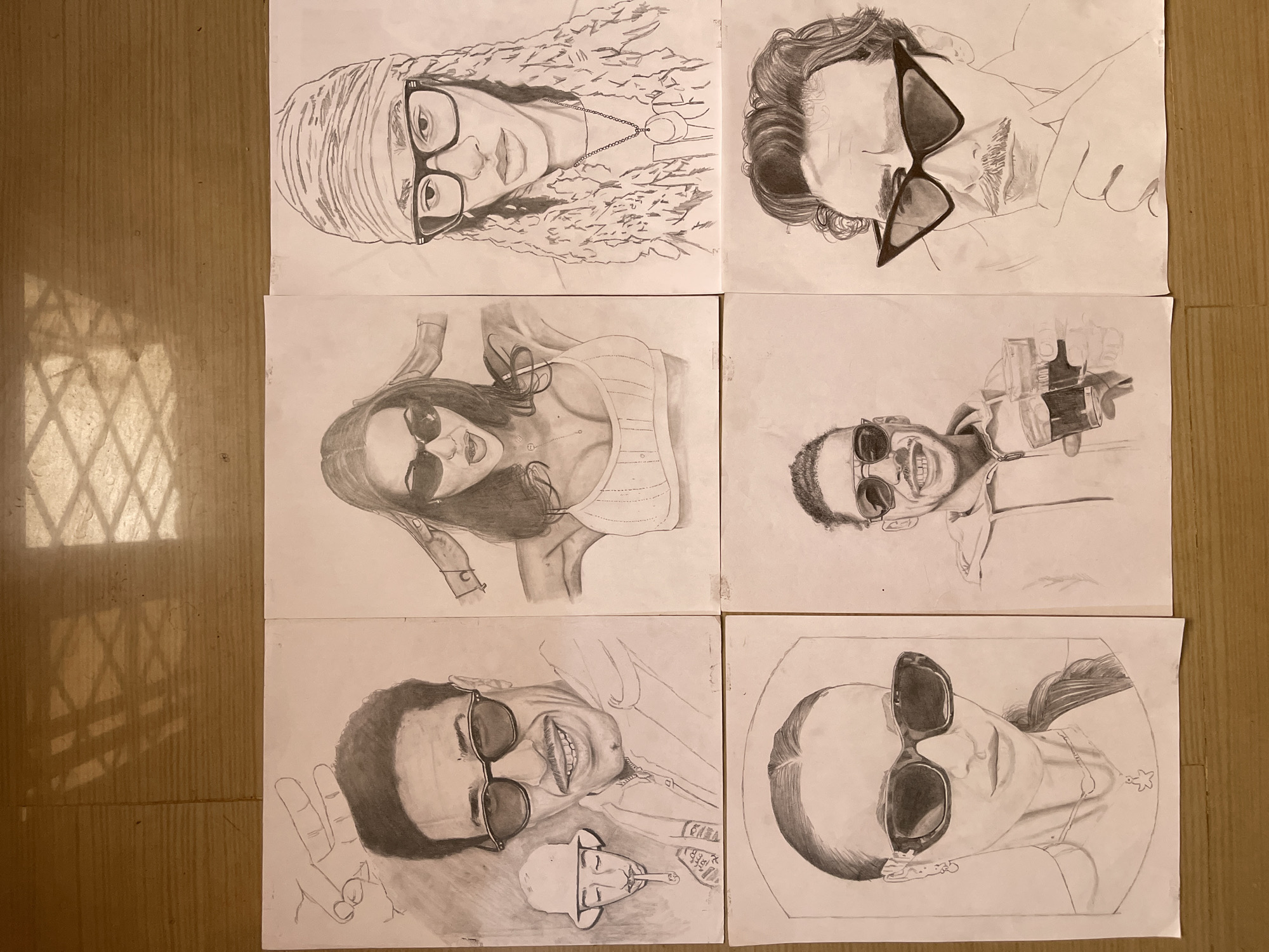 The image displays six pencil drawings on separate sheets of white paper, arranged in two rows of three on a light brown surface. The drawings are portraits of people, most wearing sunglasses or eyeglasses. The top left drawing features a person with long, curly hair, glasses, and a head covering. The top right drawing shows a person with wavy hair wearing sunglasses, partially obscured by a hand holding a drink. The middle left drawing is a portrait of a woman with sunglasses, wearing a top with straps. The middle right drawing depicts a man with sunglasses holding what appears to be a microphone. The bottom left drawing is a portrait of a man with sunglasses, smiling and holding up a peace sign. A small caricature of a man wearing a cowboy hat and smoking is visible in the lower part of the drawing. The bottom right drawing is of a woman with sunglasses and a ponytail. There is no visual information in the image that would indicate the location is Jos, Nigeria.