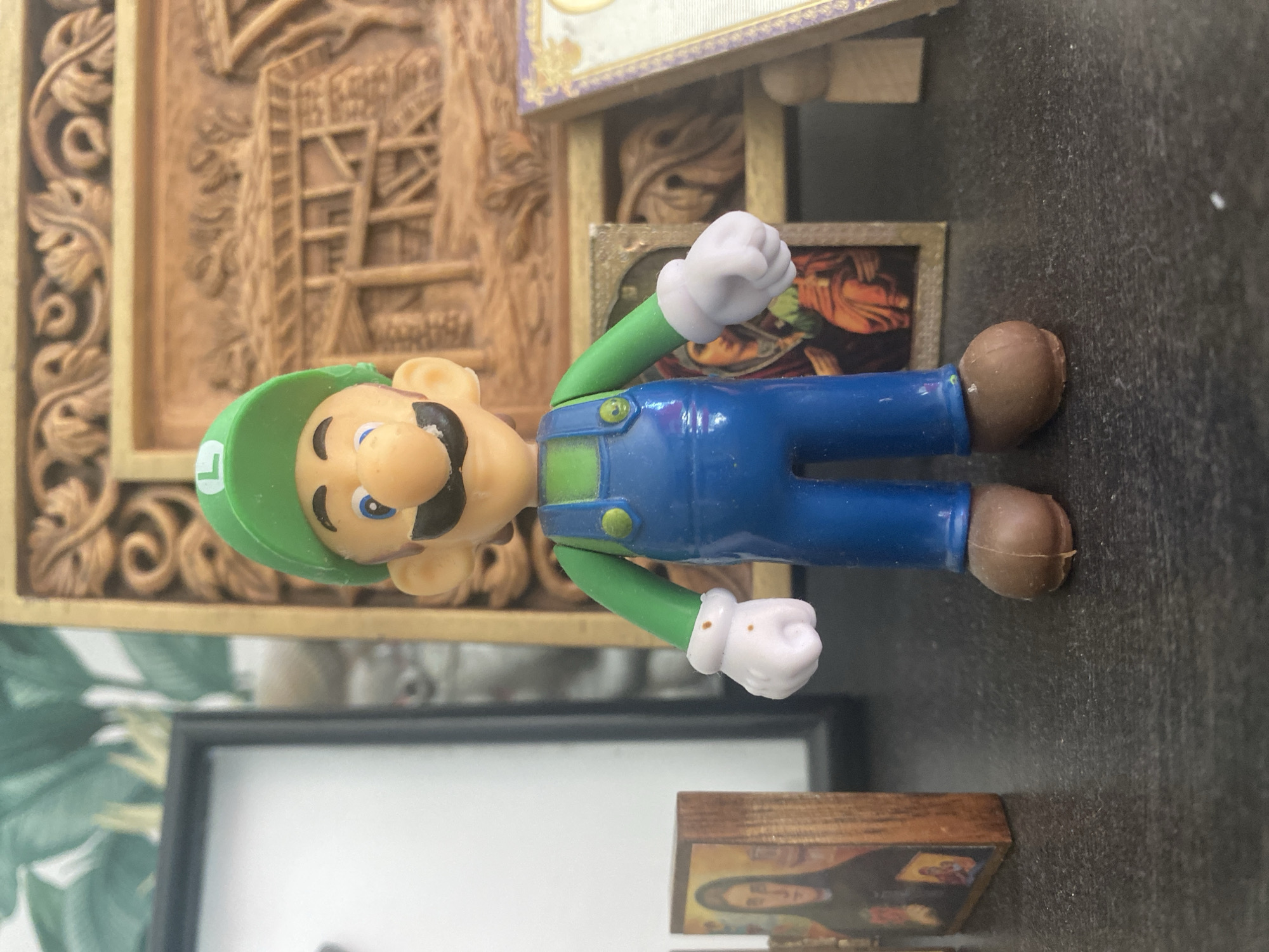 A Luigi figurine stands on a dark, textured surface. The figurine has a green hat with an "L" on it, a green shirt, blue overalls, and brown shoes. It has white gloves on its hands. Behind the figurine, there is a carved wooden panel with intricate designs. To the left of the figurine, there is a framed picture of a religious figure, and to the right, another framed religious image is visible. The background includes a portion of a black frame and a plant with green leaves. The scene is indoors and appears to be on a tabletop or shelf. The city of Blagoevgrad, Bulgaria, is not mentioned or visually represented in the image.