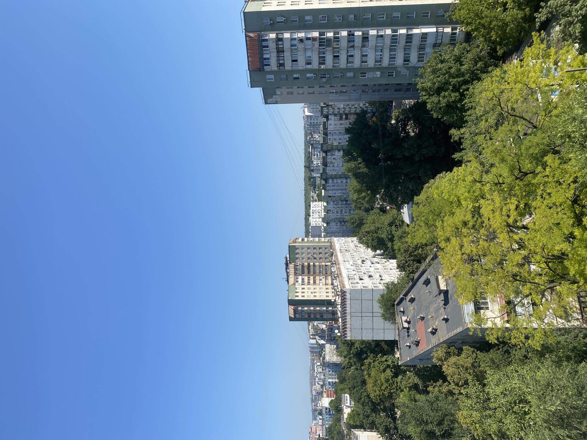 This is an aerial, eye-level, wide shot of a dense urban landscape in Chișinău, Moldova, on a clear, sunny day. The sky is a vibrant, cloudless blue, dominating the upper portion of the frame. Below the sky, a panorama of multi-story apartment buildings stretches across the horizon, interspersed with abundant green trees.
In the foreground, a flat-roofed, gray apartment building with numerous vents and pipes on its roof is visible, partially obscured by lush green trees. To its right, a tall, light green apartment building stands prominently, featuring many windows, balconies, and air conditioning units. Further into the mid-ground, a longer, multi-story building with a pale facade and green accents is present, with a distinct section of rusted, corrugated metal roofing. Other apartment buildings of varying heights and colors, some appearing older and some more modern, are scattered throughout the scene. Dense canopies of green trees and foliage fill the spaces between buildings, suggesting a well-treed city. Power lines are visible stretching across the middle distance, connecting to poles. The overall impression is one of a typical residential area in a European city. No people are visible in the image.