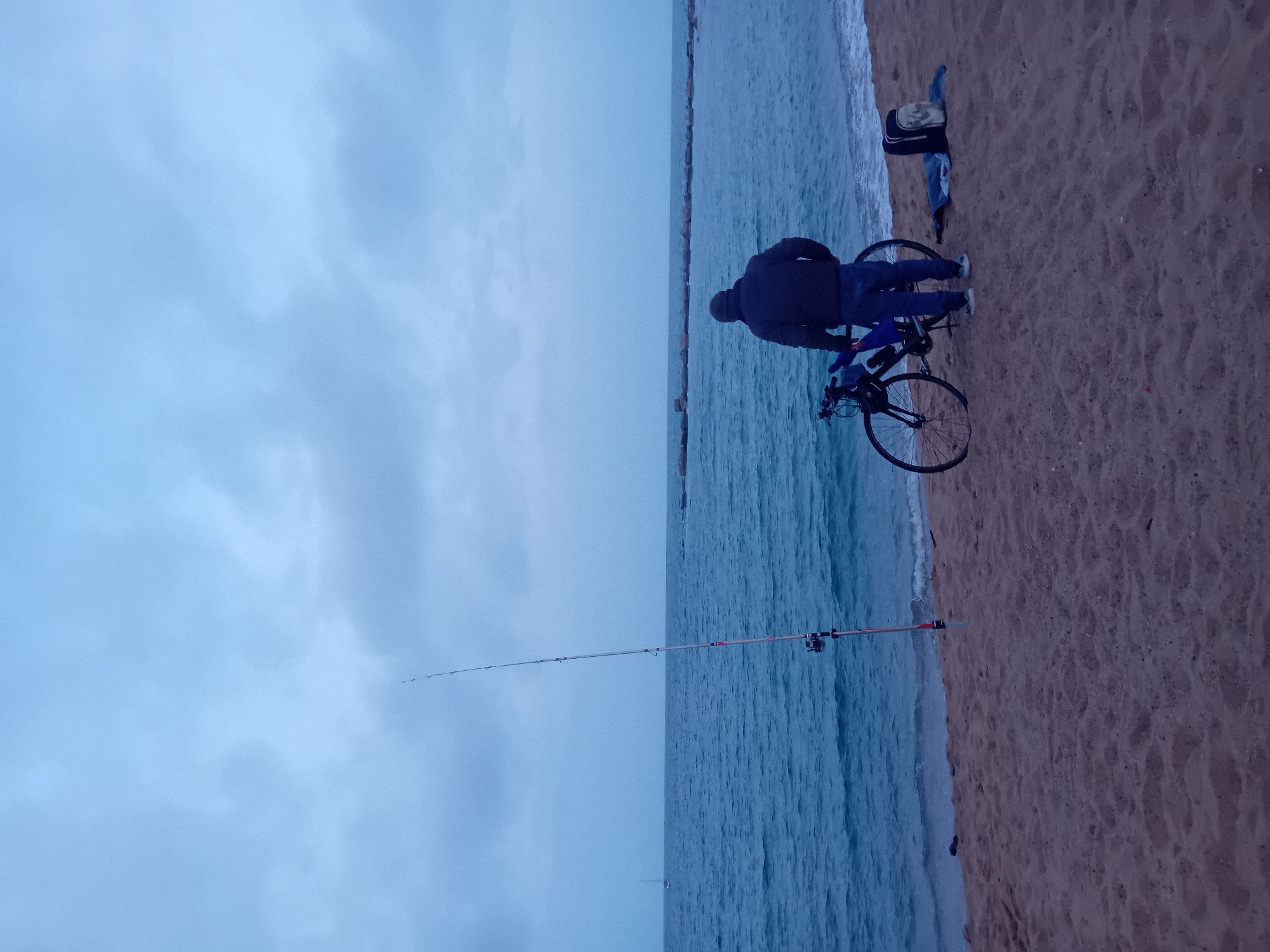 On a beach in Barcelona, Spain, a man stands near his bicycle, which is parked on the sand. He faces the sea, where a fishing rod is set up. He is wearing a dark jacket and blue jeans. A backpack and other gear are on the sand near the bike. The sky is overcast. A breakwater is visible in the distance.