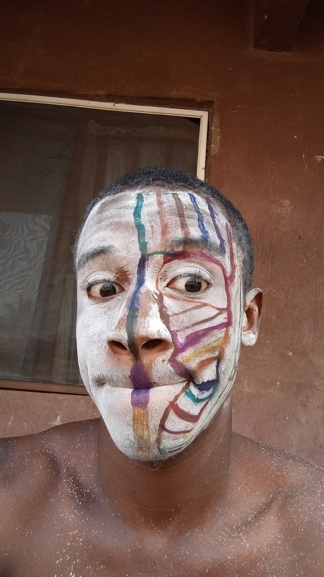 A close-up shot of a man's face with white and colorful paint. White paint covers half of his face, while the other half has vertical stripes of colors. The background reveals a window with a screen and a textured brown wall. The location appears to be Onitsha, Nigeria.