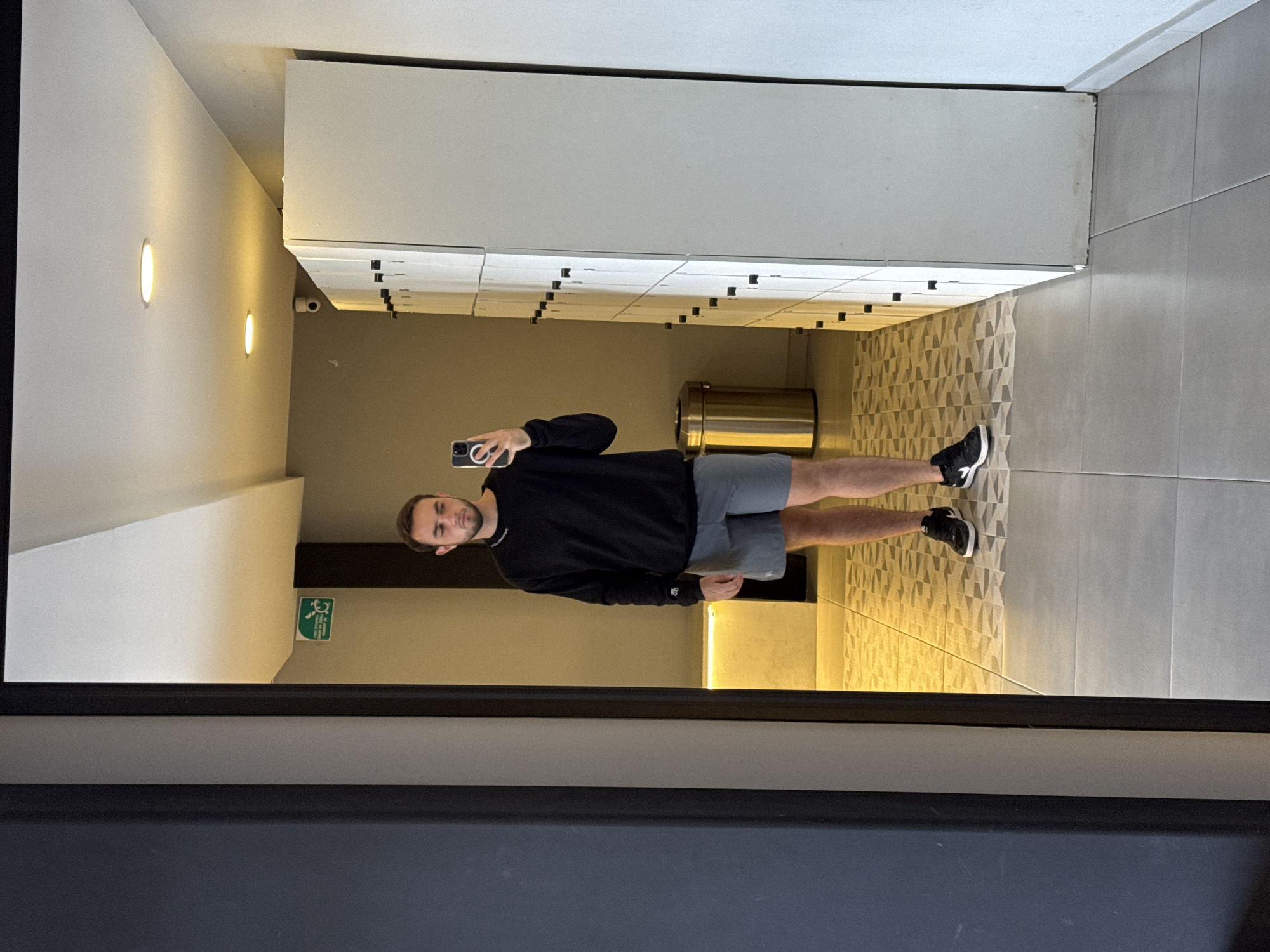 A full-length mirror reflection shows a man in a black long-sleeved shirt and grey shorts, holding a phone and taking a picture of himself. He is standing in what appears to be a gym or locker room. Behind him are rows of lockers, a cylindrical trash can, and walls with geometric-patterned tiles. Above the lockers is a white rectangular structure. The reflection suggests the setting is indoors with artificial lighting. An exit sign is visible on the wall. The location is San Bernardino Tlaxcalancingo, Mexico.
