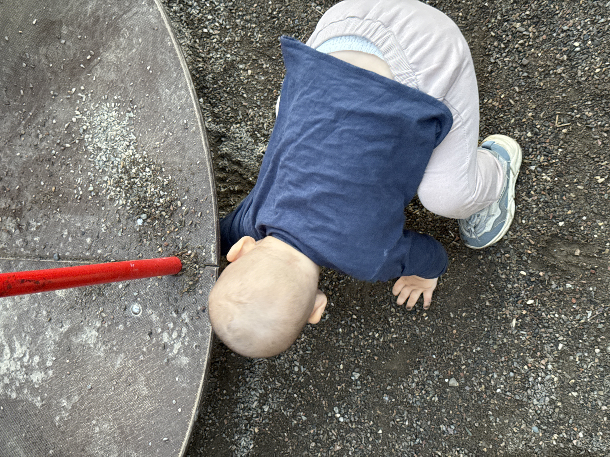 A toddler is playing on a playground in Krasnoyarsk, Russia. The child is wearing a navy blue shirt and light gray pants. They are leaning forward, with their head down, appearing to examine something on the ground. The playground surface is a mix of dark gravel and a round, textured, dark gray surface that is also covered in small pebbles. A red pole extends from the left side of the frame across the gray surface. The child's right foot, clad in a light blue and gray sneaker, is visible. The scene suggests a daytime setting, possibly overcast given the lighting. The child's posture and focus on the ground indicate engaged play.