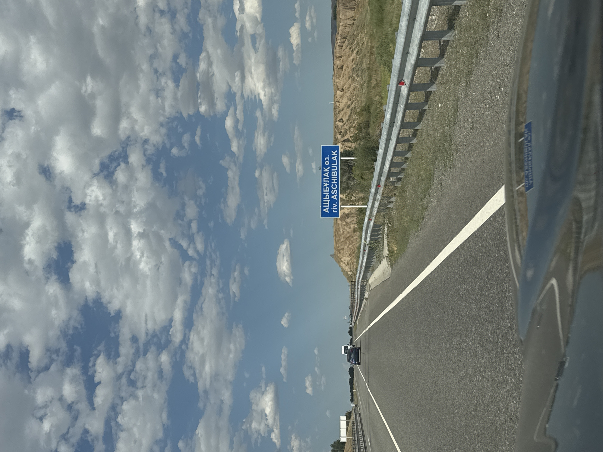The image captures a view from a moving vehicle on a highway under a partly cloudy sky. In the foreground, the hood of a dark car is visible, reflecting the sky and the road ahead. The highway has two lanes in the direction of travel, marked by white lines. A blue road sign with white text in Cyrillic and Latin alphabets is placed to the right of the road, indicating "АЩЫБҰЛАҚ өз. riv. ASCHIBULAK." This suggests the location is near the Ashibulak River, within or near the city of Koylyk, Kazakhstan. Further down the road, a dark sedan and a white van are traveling in the same direction. The right side of the highway is bordered by a metal guardrail, beyond which are dry, grassy slopes and a landscape characterized by eroded earth formations. Trees are visible in the distance on the left side of the highway.