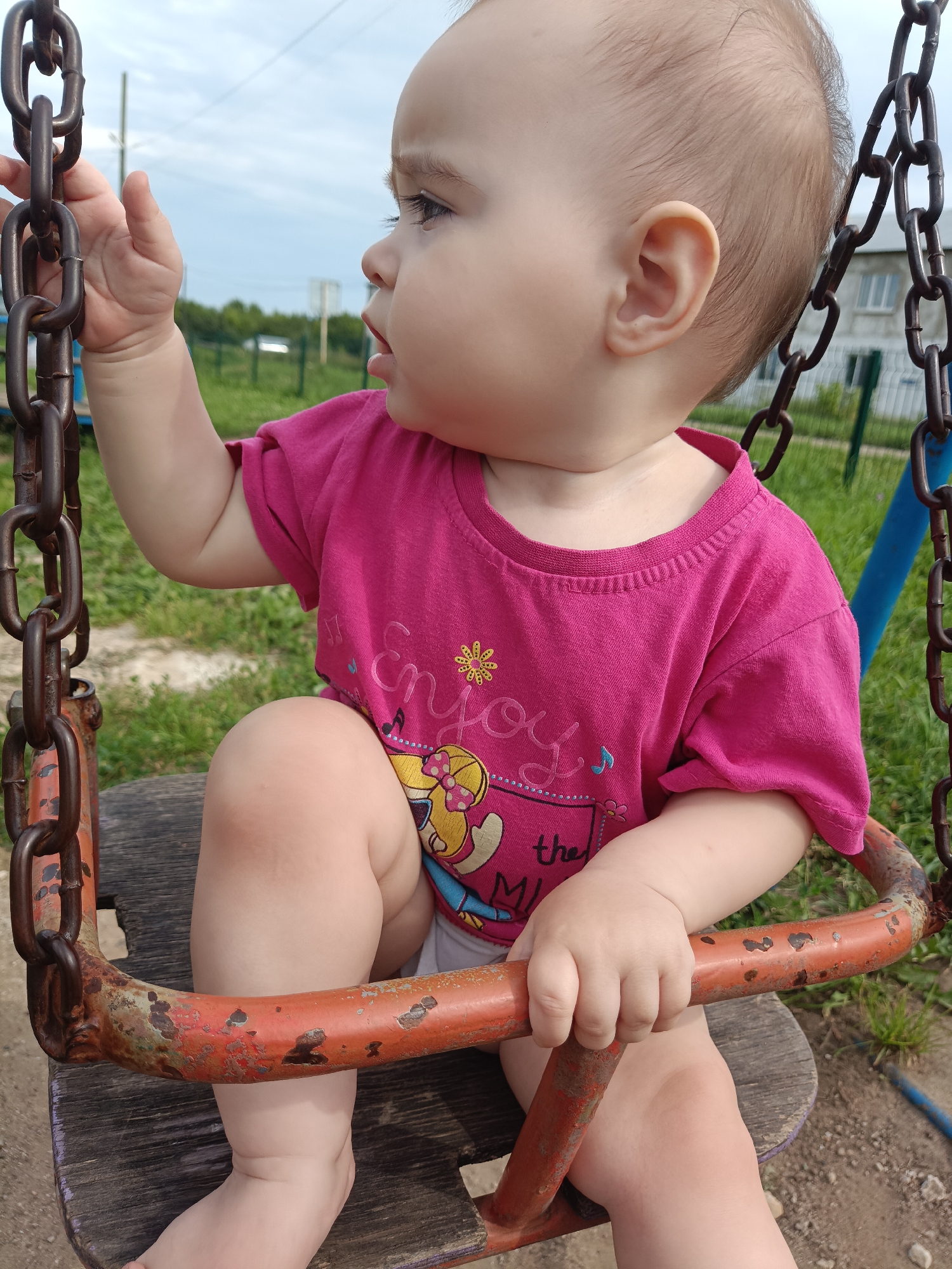 A toddler, facing left, sits on a wooden swing seat attached to rusty metal chains, which are suspended from an unseen frame. The toddler wears a bright pink t-shirt with a graphic print. Their right hand is extended upwards, touching one of the chains. The background depicts an outdoor environment with green grass, a fence, trees, and a building with windows under a cloudy sky.