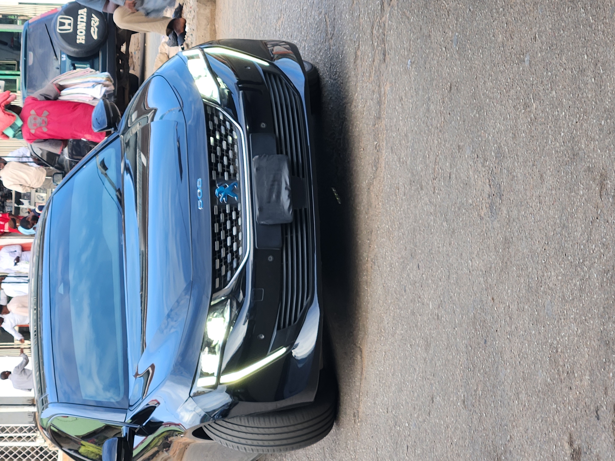 The image displays an outdoor street scene during bright daylight. The main subject is the front left side of a dark blue/black Peugeot 508 sedan, parked on an asphalt or concrete road. The car features modern LED headlights and daytime running lights, and its "508" badge and Peugeot lion logo are visible on the front grille.
In the background, an older model dark-colored Honda CR-V is parked, identifiable by the "HONDA CR-V" text on its rear spare tire cover. Bundles of clothes or fabric are stacked on the back of the CR-V. Several people are visible in the background: one person wears a distinctive red top with a white design, while others are seen in light-colored clothing, standing or moving around.
The setting appears to be a busy urban or semi-urban street, consistent with daily life and commerce. The bright sunlight suggests a clear day, and reflections on the car's surface indicate a sunny environment. No specific event is taking place, but rather a snapshot of an active street. The visible vehicles and general atmosphere align with an urban setting such as Jos, Nigeria.