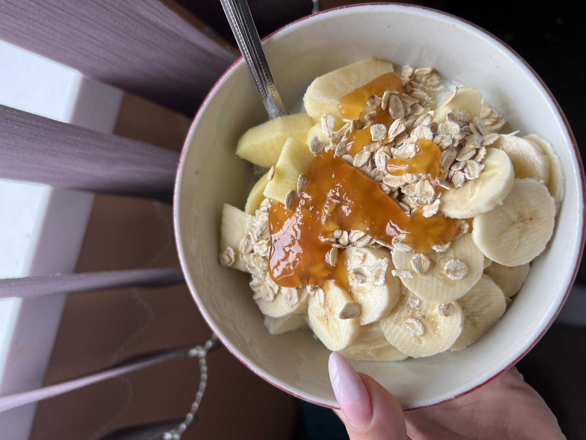 A top-down, close-up shot features a white bowl with a red rim, held in a person's hand, containing a breakfast dish. The dish consists of sliced bananas and apples, sprinkled with oats, and drizzled with a viscous, amber-colored substance resembling jam or honey. A silver spoon is partially visible, inserted into the food. The person's hand holding the bowl has a long, pink-hued fingernail. The background includes partially visible blinds, suggesting an indoor setting. The location cannot be determined to be in Plovdiv, Bulgaria, with the available information.