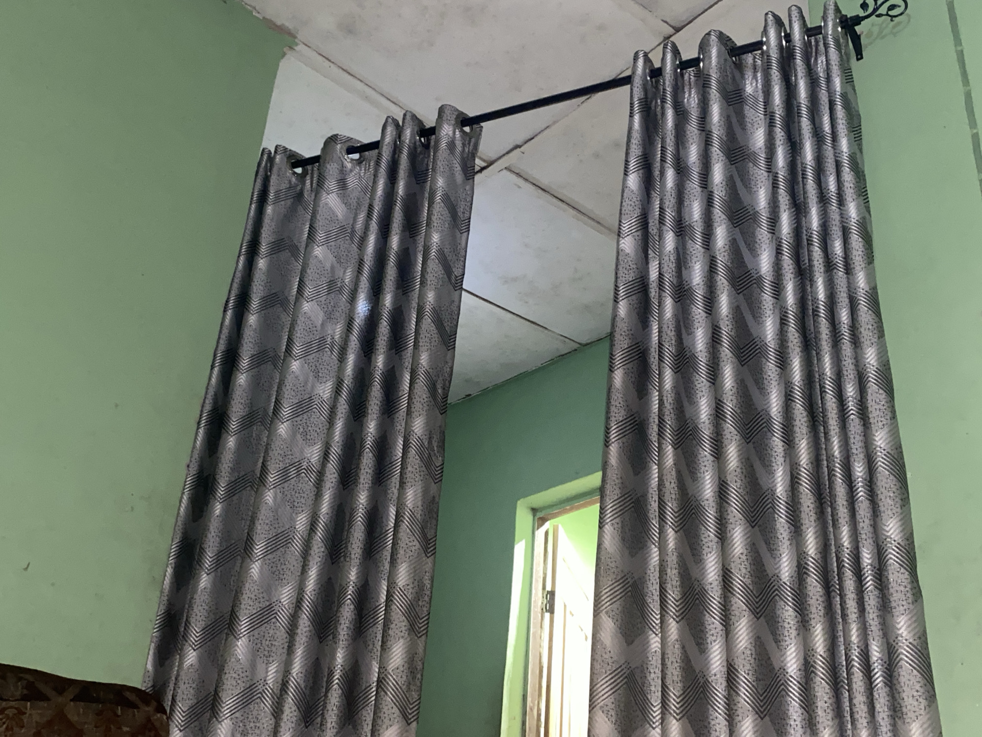 The image shows a pair of gray patterned curtains hanging from a black rod. The curtains are partially drawn, revealing a glimpse of a door and a light green wall. The walls in the room are painted in light green, and the ceiling appears to be made of white tiles or panels. The location appears to be indoors.