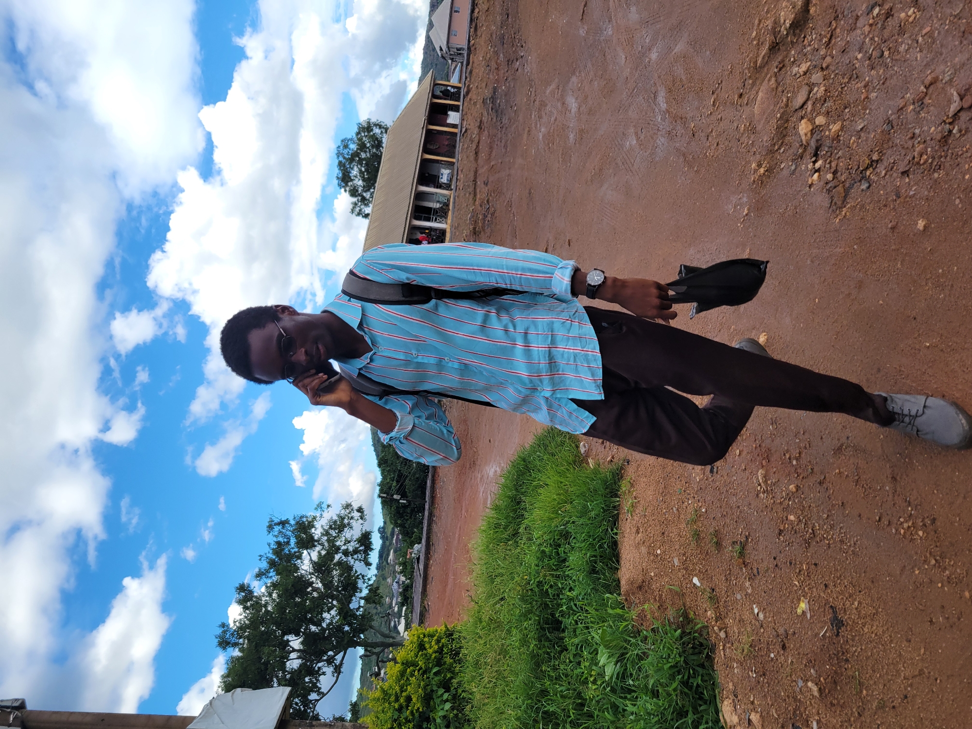 A full-body shot captures a Black male wearing glasses, a blue and white striped button-up shirt, brown pants, and gray shoes. He is walking away from the camera, looking down at his phone as he speaks into it. He is also carrying a black bag in his right hand. A backpack is visible on his shoulders.
The man is walking on a dirt road with patches of wet mud and scattered pebbles. To his left, there is a dense patch of bright green grass. In the background, a building with a yellow roof and open-air seating is visible, along with other structures further in the distance. The sky above is a vibrant blue with scattered white clouds. The overall environment suggests an outdoor location in a rural or semi-urban area, possibly in Jos, Nigeria, given the reddish-brown soil and the style of buildings.