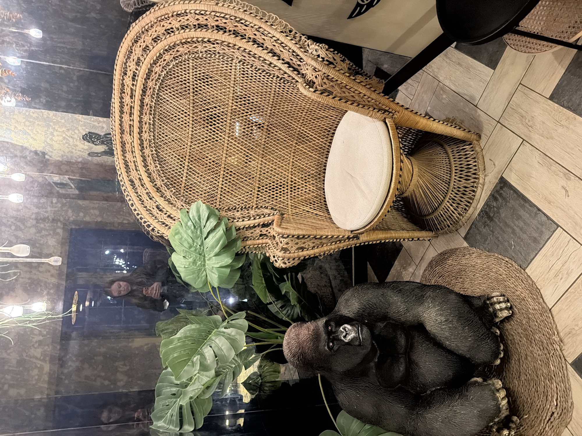The image shows a high-angle shot of a lounge area with eclectic decor. A prominent wicker peacock chair with a white cushion sits on a patterned floor of light and dark tiles. A reflective black surface, possibly a table or a polished floor, displays a reflection of a person holding a red object and the surrounding environment, including a wall-mounted lighting fixture. Lush green artificial monstera leaves cascade over the edge of the black surface, partially obscuring the reflection. At the bottom right, a lifelike sculpture of a gorilla rests on another woven base. The scene is indoors. This location could be within a bar/restaurant called 'El Barco', located in Varna, Bulgaria.