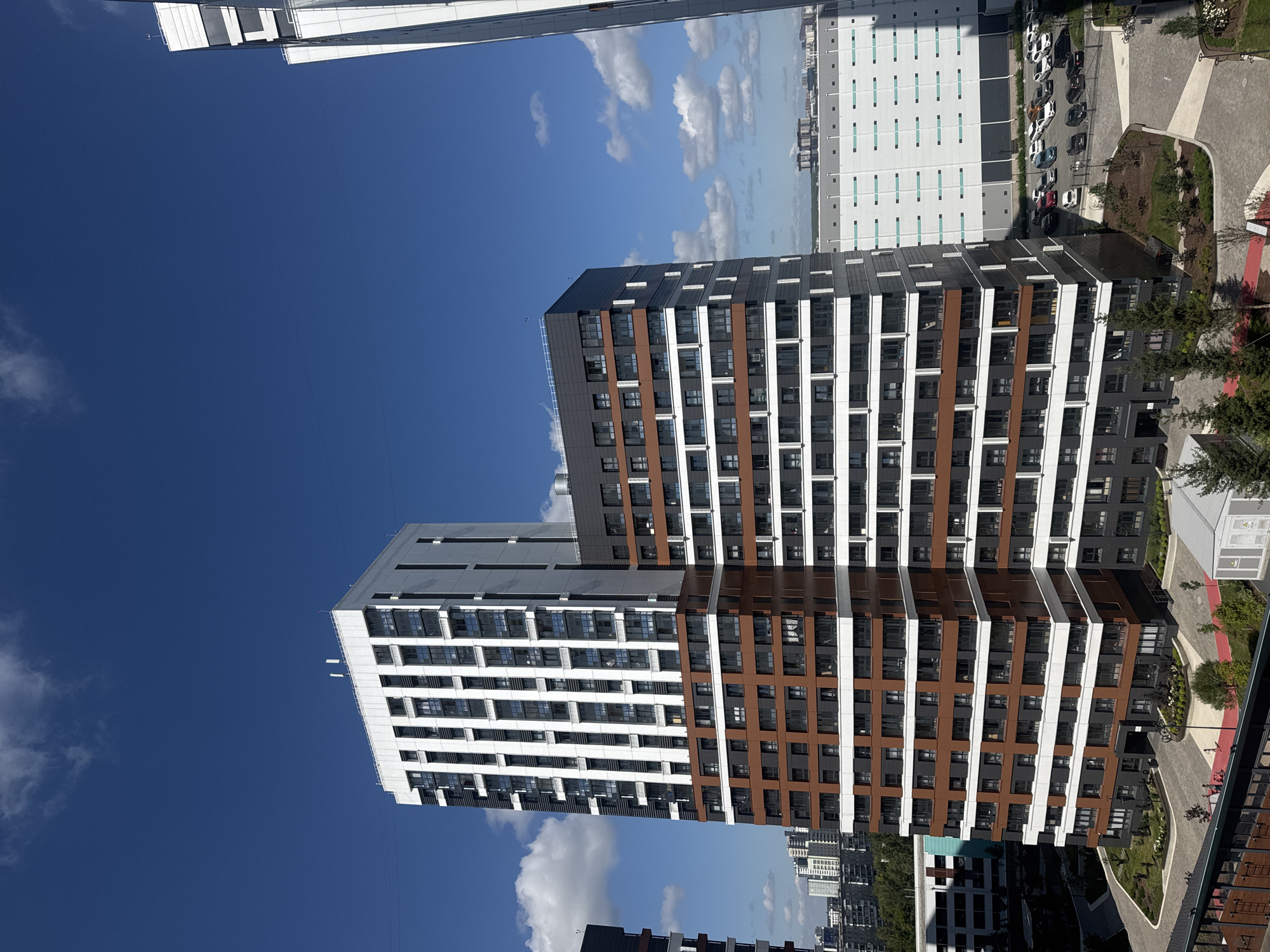 The image captures a modern residential complex in Novosibirsk, Russia, under a clear blue sky dotted with white clouds. The dominant structures are several tall, multi-story buildings with distinct architectural features. The main building in the foreground is a high-rise with a white top section featuring numerous vertical windows and balconies, juxtaposed with a lower section clad in brown and dark grey panels. Adjacent to it is another building, also tall, with a similar design, incorporating white, dark grey, and brown elements.
The surrounding environment includes a paved courtyard area with landscaping, featuring trees, bushes, and pathways. A red carpet-like path is visible leading towards one of the building entrances. A parking lot filled with various cars is situated behind the buildings, with a large, industrial-looking white building in the background, possibly a warehouse or commercial facility. The scene suggests a contemporary urban development.