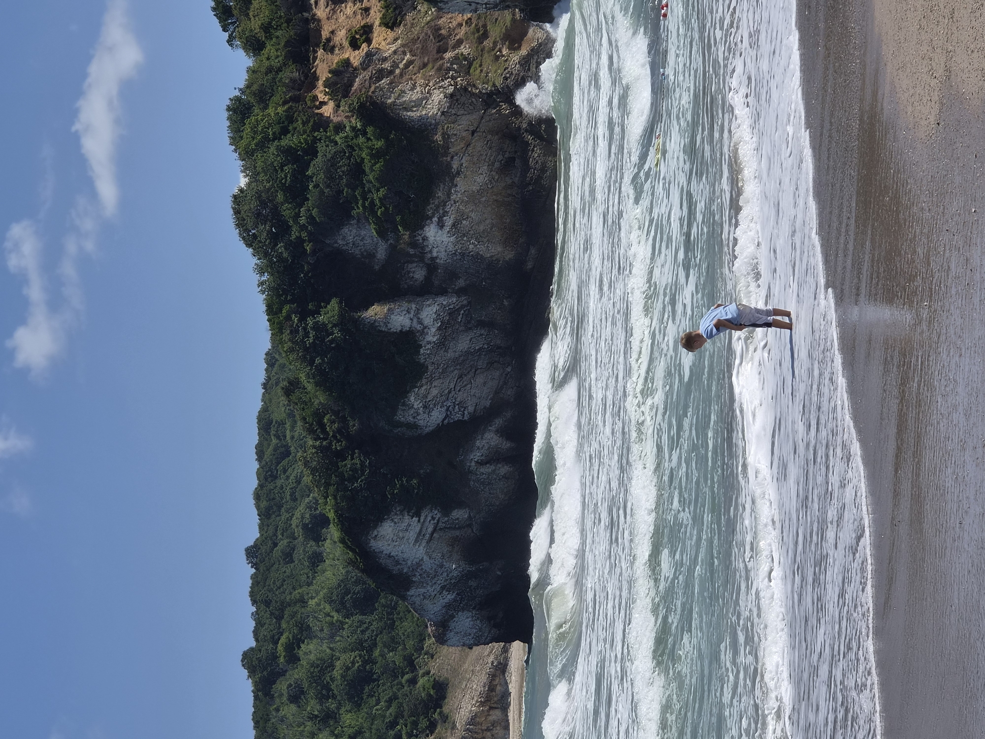 A lone young boy, wearing a light blue t-shirt and grey shorts, stands with his feet in the shallow water of a beach. The boy is facing away from the camera, looking down towards the water. He appears to be contemplating something in the surf. The beach is composed of sand and the water is a vibrant turquoise color, with moderate waves breaking gently onto the shore. In the background, a large, imposing rock formation with a dense, green forest covering its top rises from the sea. The sky is a clear blue with a few wispy white clouds scattered across it. This scene is characteristic of the Black Sea coast near Şile, Turkey, a region known for its dramatic cliffs and beautiful beaches.