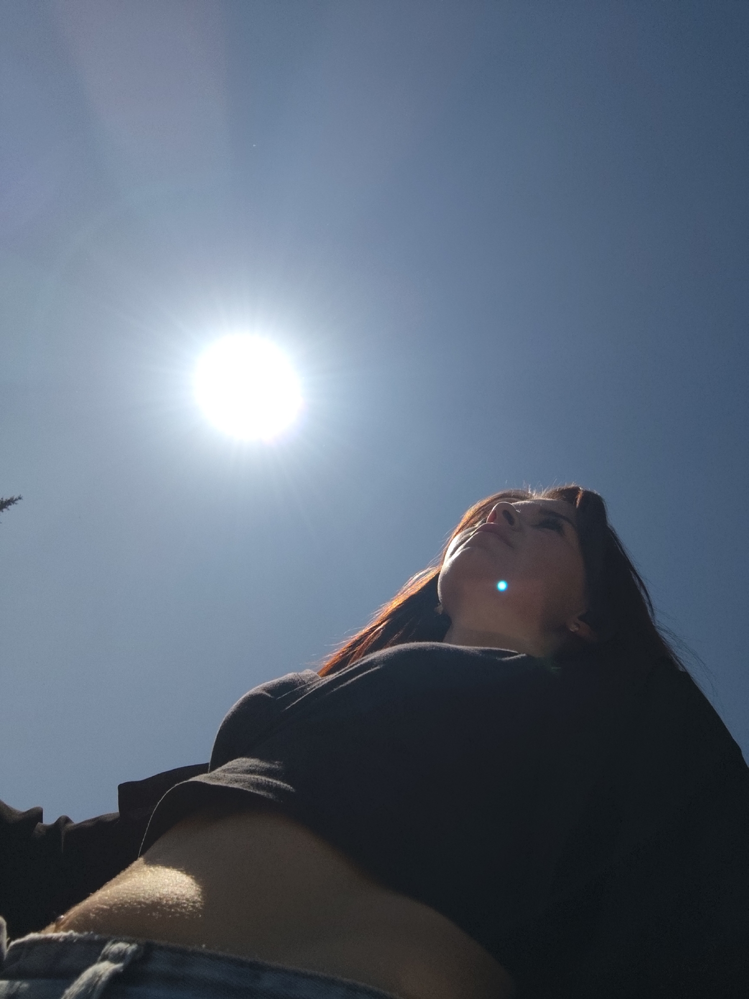 A low-angle, full-body outdoor shot features a female facing upward toward the sun. The location is not identifiable as Chaikovsky, Russia. The woman has dark hair with reddish highlights, and she is wearing a dark-colored shirt, exposing a portion of her abdomen. The sky is blue with a bright sun in the upper left quadrant of the frame. The lower part of her body is cut off by the frame, showing a portion of her jeans.