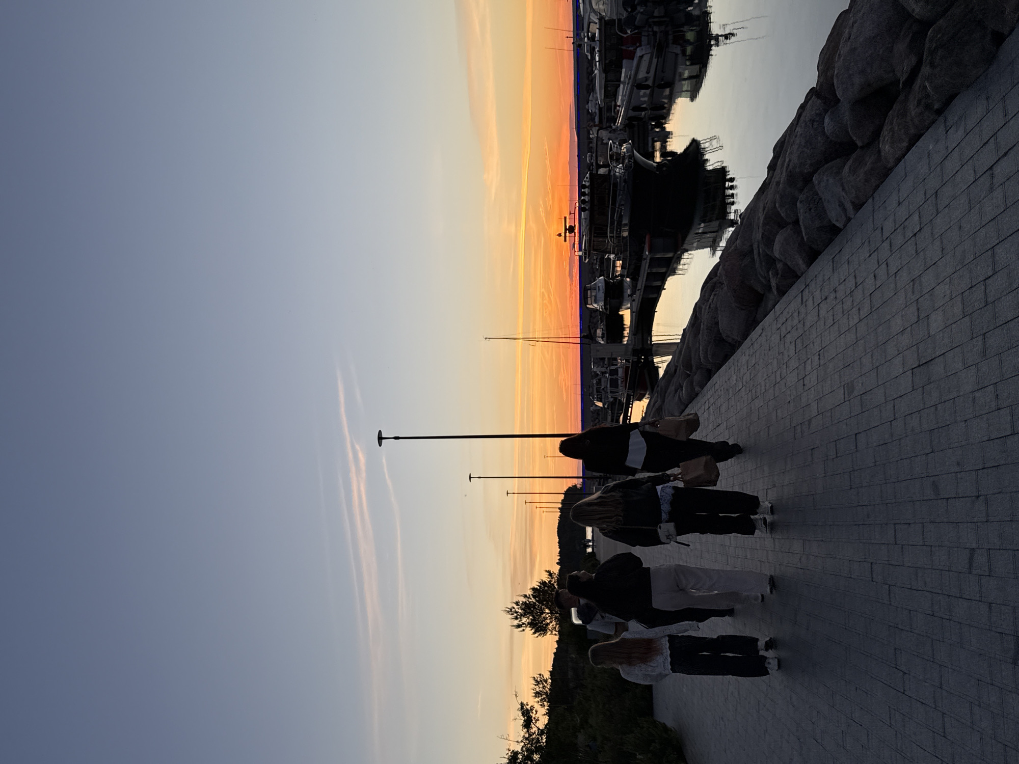 The image depicts a group of five individuals, appearing to be young adults, walking away from the camera on a paved walkway alongside a harbor. The setting is likely Tallinn, Estonia, given the presence of boats and the twilight sky. The walkway is constructed of dark grey rectangular pavers. To the right of the walkway, a rocky embankment lines the water, where several boats are docked. The water reflects the twilight sky, which is a gradient of pale blue at the top to vibrant orange and yellow near the horizon. A few thin clouds are visible in the sky, tinged with orange from the setting sun. The individuals are dressed in casual attire. Two of them are carrying shopping bags. In the background, trees and more distant lights are visible. The overall mood is peaceful and serene, characteristic of an evening scene at a harbor.
