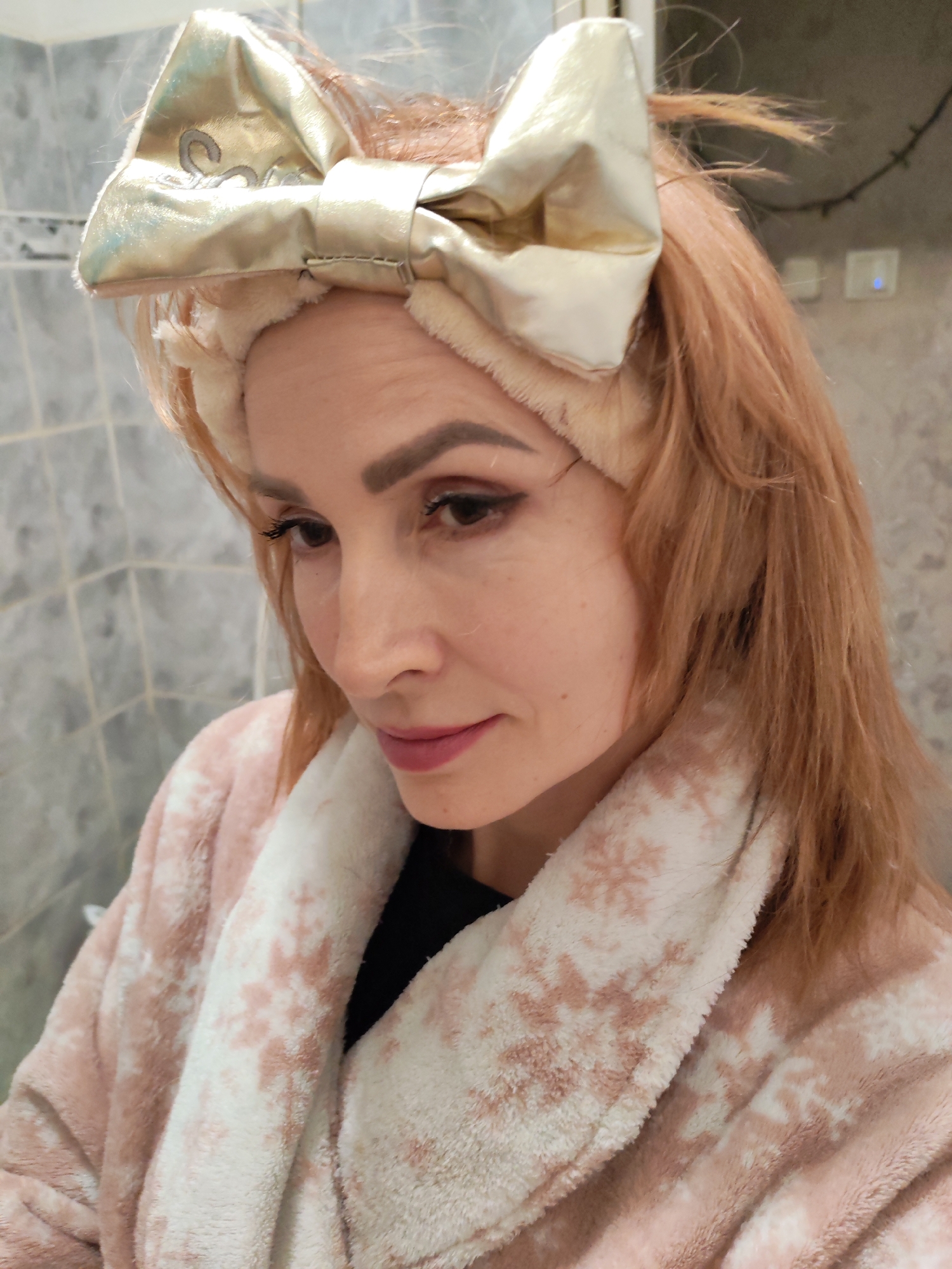 A woman is shown in a close-up portrait, likely indoors. She has light brown hair, light skin, and is wearing a gold bow headband. She is also wearing a pink and white robe. The background appears to be a tiled wall. The location is Chaikovsky, Russia.