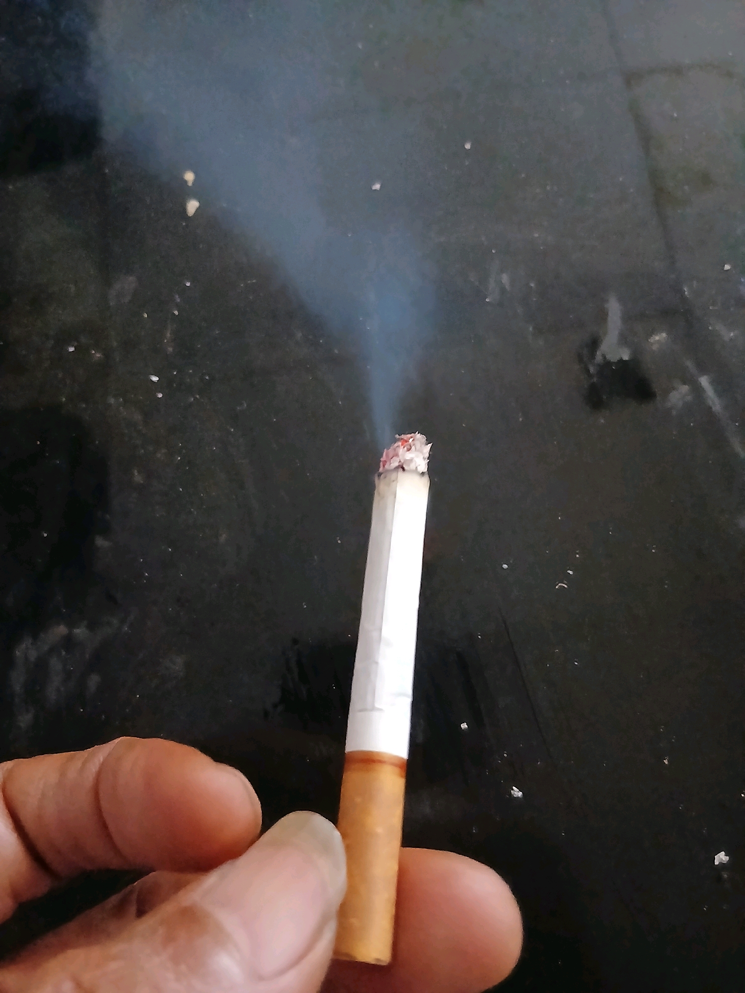 In Kademangan Kulon, Indonesia, a close-up, slightly angled shot depicts a person's hand holding a lit cigarette. The cigarette, with its glowing ember and wisps of smoke, is positioned against a dark, speckled surface. The hand, with visible fingers and nails, grips the cigarette from below. The background is a dark, textured surface, possibly a table, with small specks of debris scattered across it. The lighting suggests an indoor environment, with the smoke catching the light and creating a hazy effect.