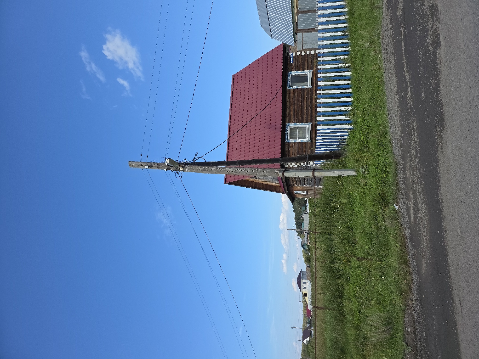 The image depicts a rustic wooden house with a red metal roof in the village of Khalitova, Russia. The house is situated behind a fence made of vertical blue and white planks. A wooden utility pole with electrical wires stands in front of the house, to the left of the fence. The scene is set against a clear blue sky with a few wispy white clouds. In the background, there are other houses and trees, suggesting a rural or semi-rural environment. The ground in the foreground appears to be a dirt or gravel road. There are no people visible in the image.