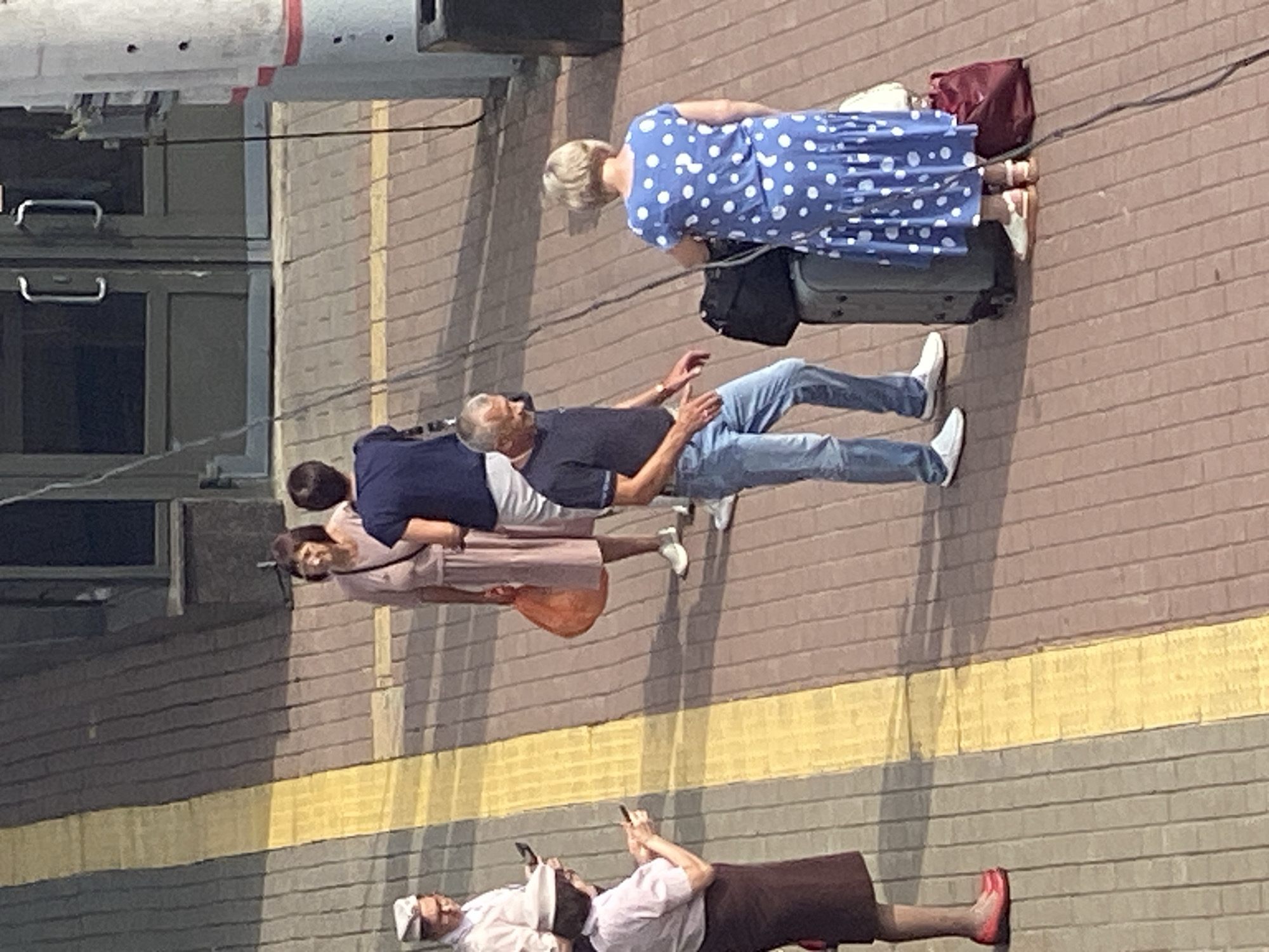 The image depicts an outdoor scene, likely a train station platform in Novokuznetsk, Russia, given the context provided. Several individuals are visible, most standing with luggage.
On the left, two women are present. One, facing away from the camera, wears a pink top and a dark skirt, holding a mobile device. Another woman, further back and facing left, is wearing a pink dress and carrying an orange bag. A man in a white cap and uniform stands to the far left, also holding a mobile device.
In the center of the frame, a man in a dark blue shirt and jeans stands with his hands gesturing slightly, facing a woman in a blue polka-dot dress. The woman is holding onto a suitcase. Behind them, another dark bag rests on the platform.
The platform is paved with brick-like tiles, with a distinct yellow line running along the edge of the walking area. The background shows a building with large glass doors and a concrete pillar. The lighting suggests it is daytime, with shadows cast by the people and objects.