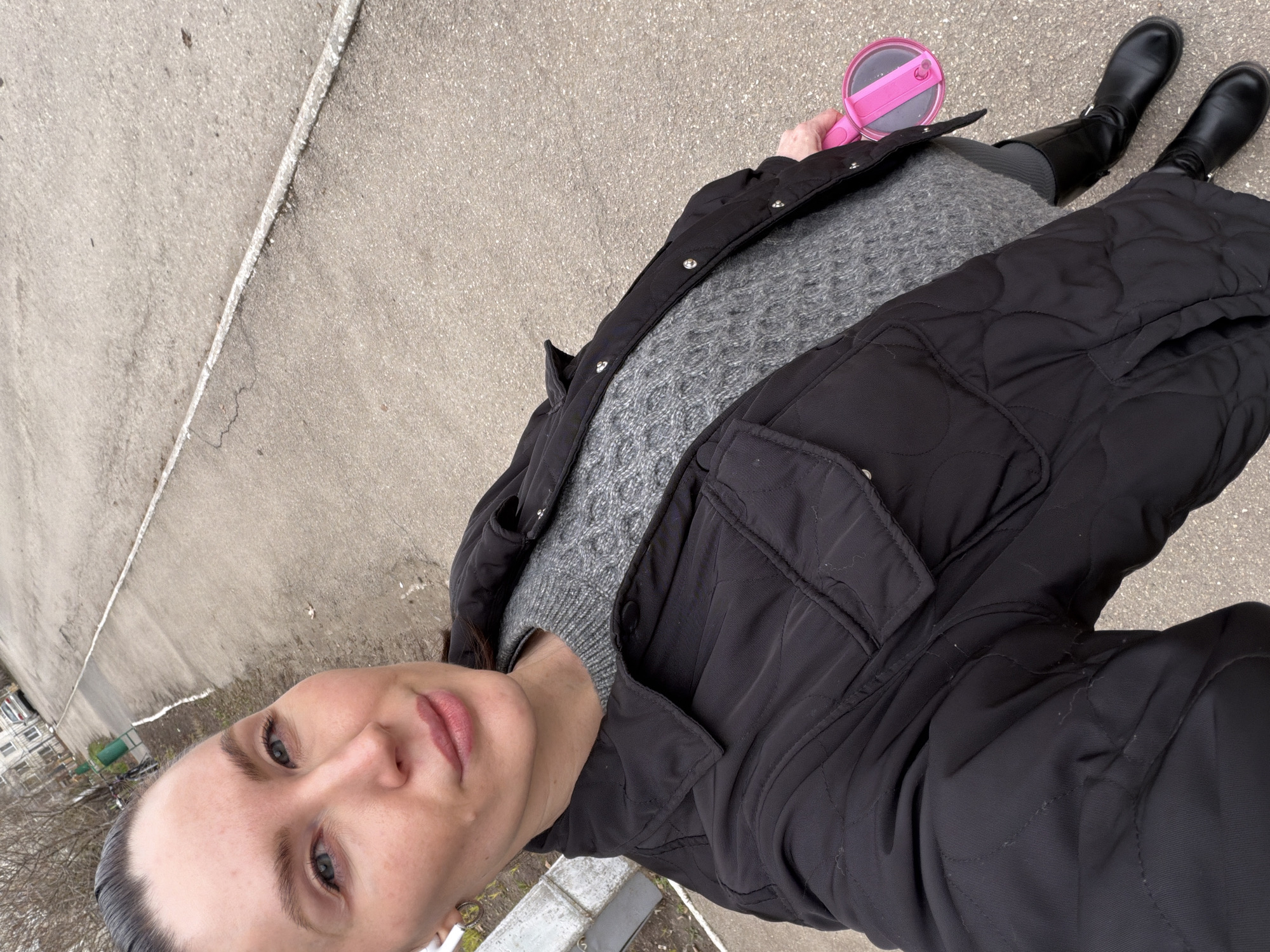 A self-portrait captures a woman from an overhead, slightly angled perspective, focusing on her upper body and legs. She has dark hair pulled back, light-colored eyes, and wears light makeup, including pinkish lips. Her attire consists of a grey, textured sweater layered under a black quilted jacket, and she is wearing black boots. In her right hand, she holds a distinctive pink object with a clear circular top, resembling a small mirror or a child's toy.
The setting is outdoors, with a concrete or asphalt ground serving as the primary background, featuring a faded white line running diagonally across it. In the distance, glimpses of bare trees and urban buildings typical of a city environment are visible. The scene is located in Perm, Russia. The overcast sky provides diffused lighting, and the woman's warm clothing along with the leafless trees indicate a cool season like late autumn, winter, or early spring.
The woman is engaged in taking a selfie, looking calmly towards the camera. No visible text is present in the image.