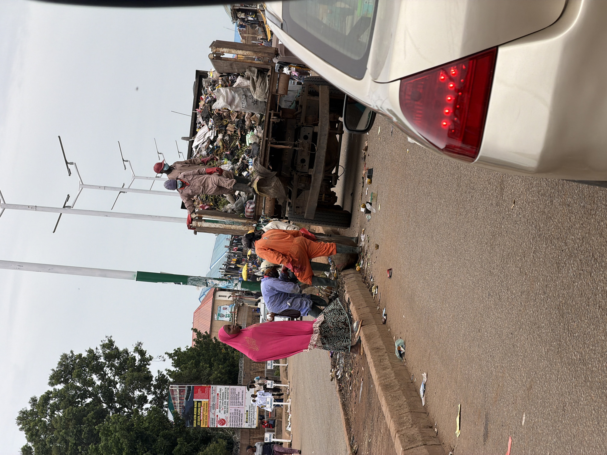 The image depicts a street scene in Bukuru, Nigeria, with a garbage truck overflowing with waste. Several people, seemingly workers, are tending to the garbage. One individual in an orange jumpsuit and another in a light blue outfit are close to the truck, while a person in a pink hijab stands nearby. The scene appears to be in daylight under an overcast sky. The road is paved, and a concrete curb runs alongside it. Debris and trash are scattered on the ground and along the curb. In the background, buildings and trees are visible, along with streetlights. A poster with text, possibly an advertisement or announcement, is prominently displayed near some trees, reading "BUKURU ULTRA MODERN MARKET JOS SOUTH, PLATEAU STATE" and providing contact numbers. Several "NO PARKING" signs are also visible. The overall impression is one of an active, albeit somewhat messy, urban environment where waste management is taking place.