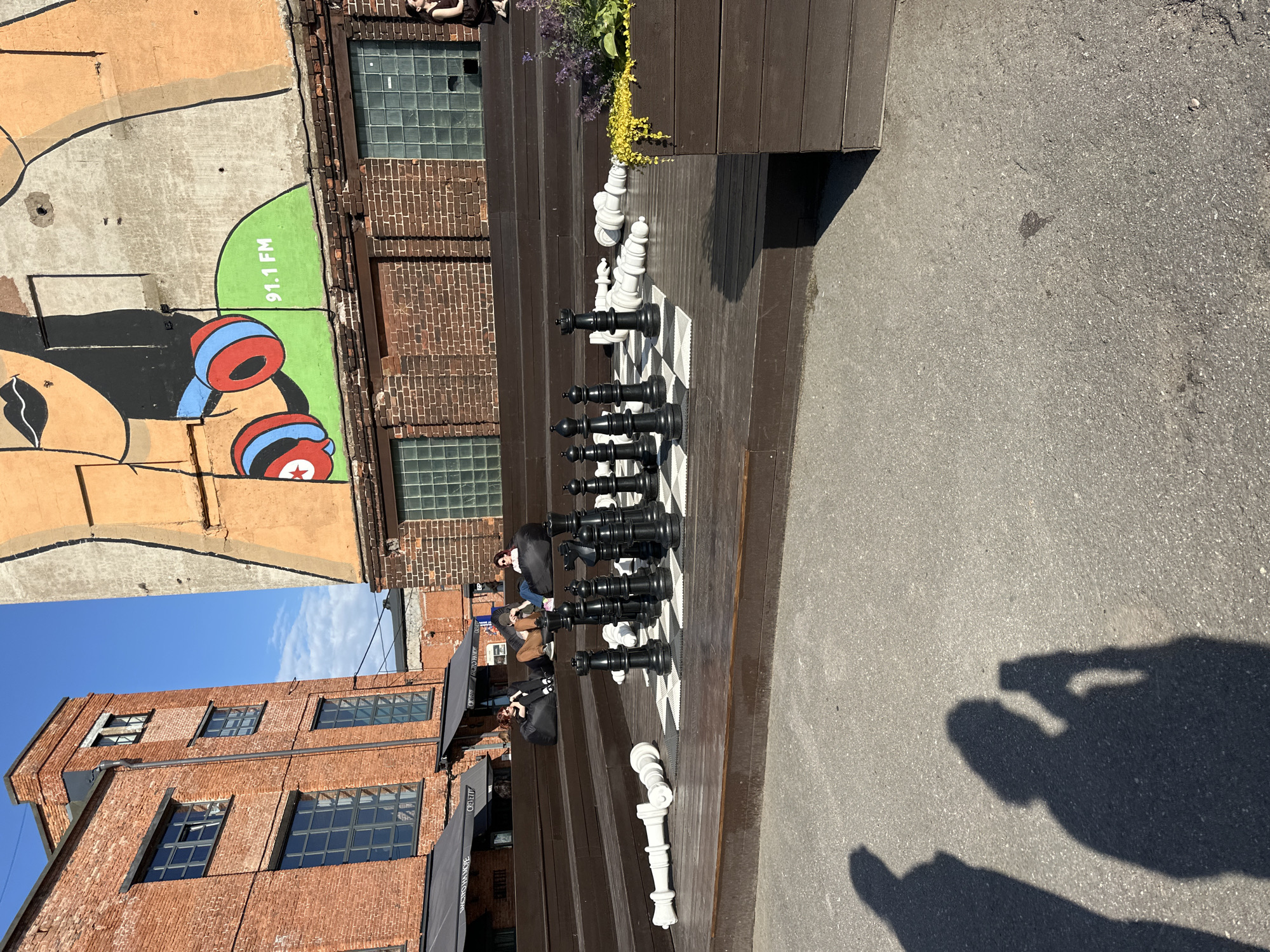 The image depicts an outdoor seating area with a giant chess set at a location in Saint Petersburg, Russia. In the foreground, a large chess board is set up with black and white pieces, some of which are fallen. Behind the chess set, several people are seated on beanbag chairs on tiered wooden steps, enjoying the sunny day. The background features red brick buildings and a mural on a wall, typical of industrial or artistic urban spaces. The shadows of two people taking a selfie are visible on the asphalt in the lower left corner.