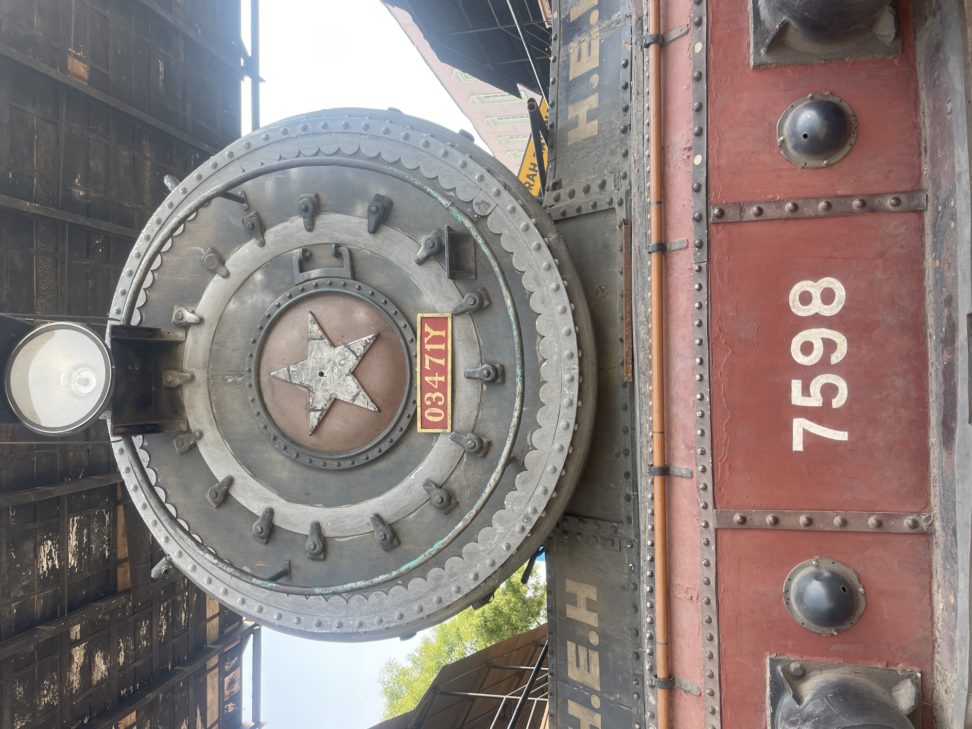 The image shows a close-up of a portion of a vintage steam locomotive. The front face of the boiler is visible, featuring a large circular door with a raised star emblem in the center and the number "0347IY" on a small plate. A headlight is mounted on the left side of the boiler. Adjacent to the boiler is a maroon-colored side panel with the number "7598" painted in white. The environment includes parts of a building with a dark facade. The location is implied to be Anajpur, India.