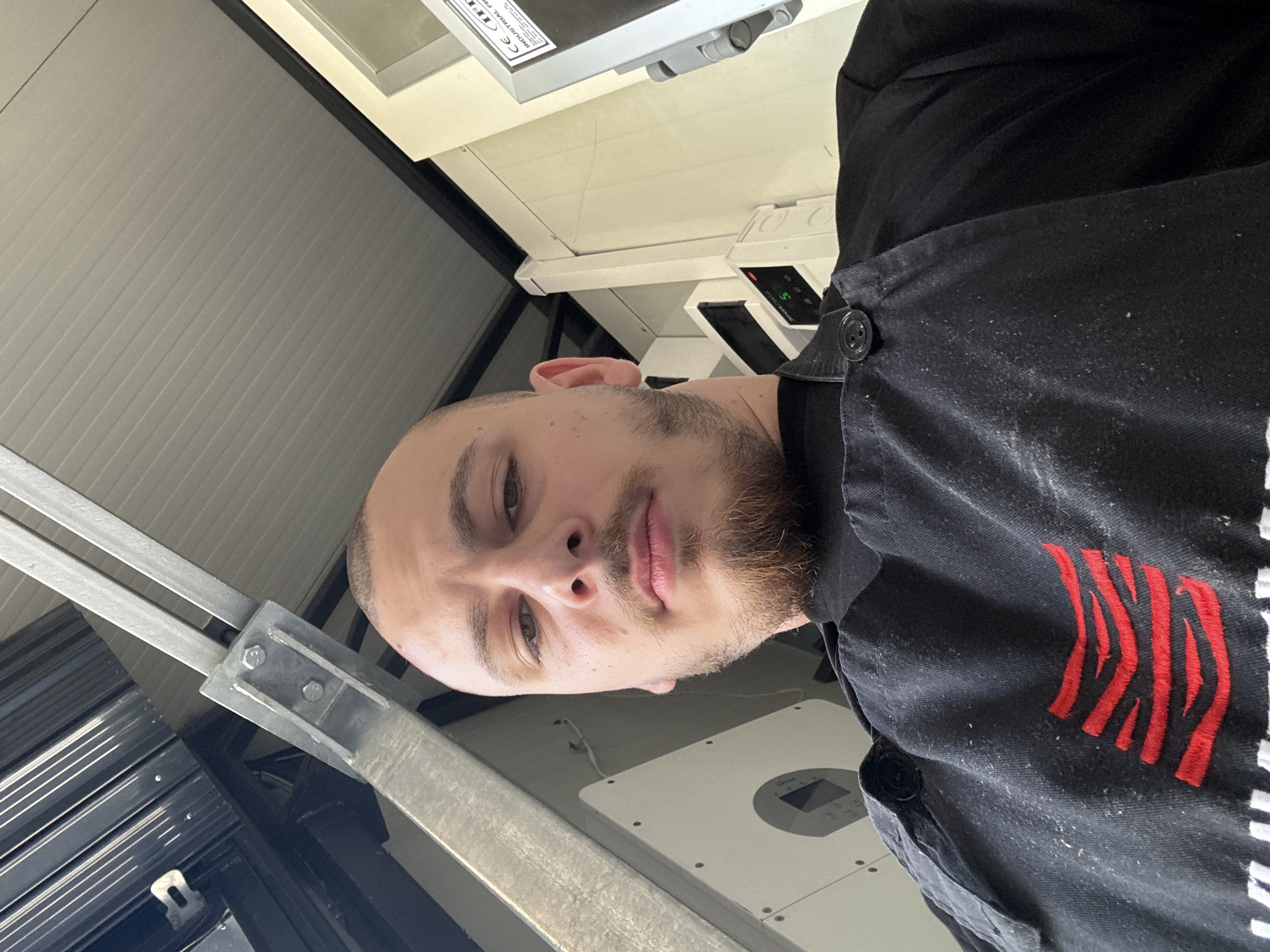 A man with short hair and a beard is in what appears to be an industrial or commercial setting. He is wearing a dark-colored work shirt or jacket with red markings on the front. Behind him, there is equipment that suggests a possible manufacturing or engineering environment. There is a white box-like object with a circular design and several screws visible. The man is looking directly at the camera, and the image is slightly tilted. There is metal framing in the background with a light-colored, corrugated metal wall. It is not possible to determine the location in Stara Zagora, Bulgaria, from the image.