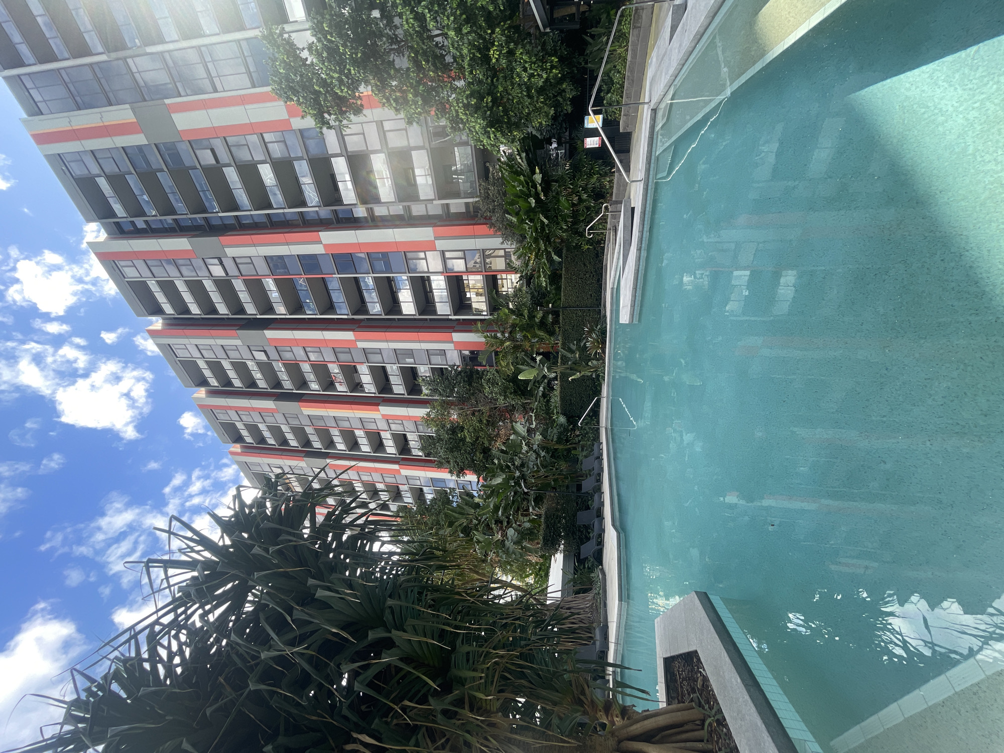 The image shows a swimming pool in front of a modern apartment building in Brisbane, Australia. The building is tall and has a grey facade with red and orange vertical stripes. The pool is a bright turquoise color and has a tiled bottom. There are lounge chairs around the pool, as well as lush greenery. The sky is blue with some white clouds. The overall impression is one of a relaxing and luxurious urban oasis.